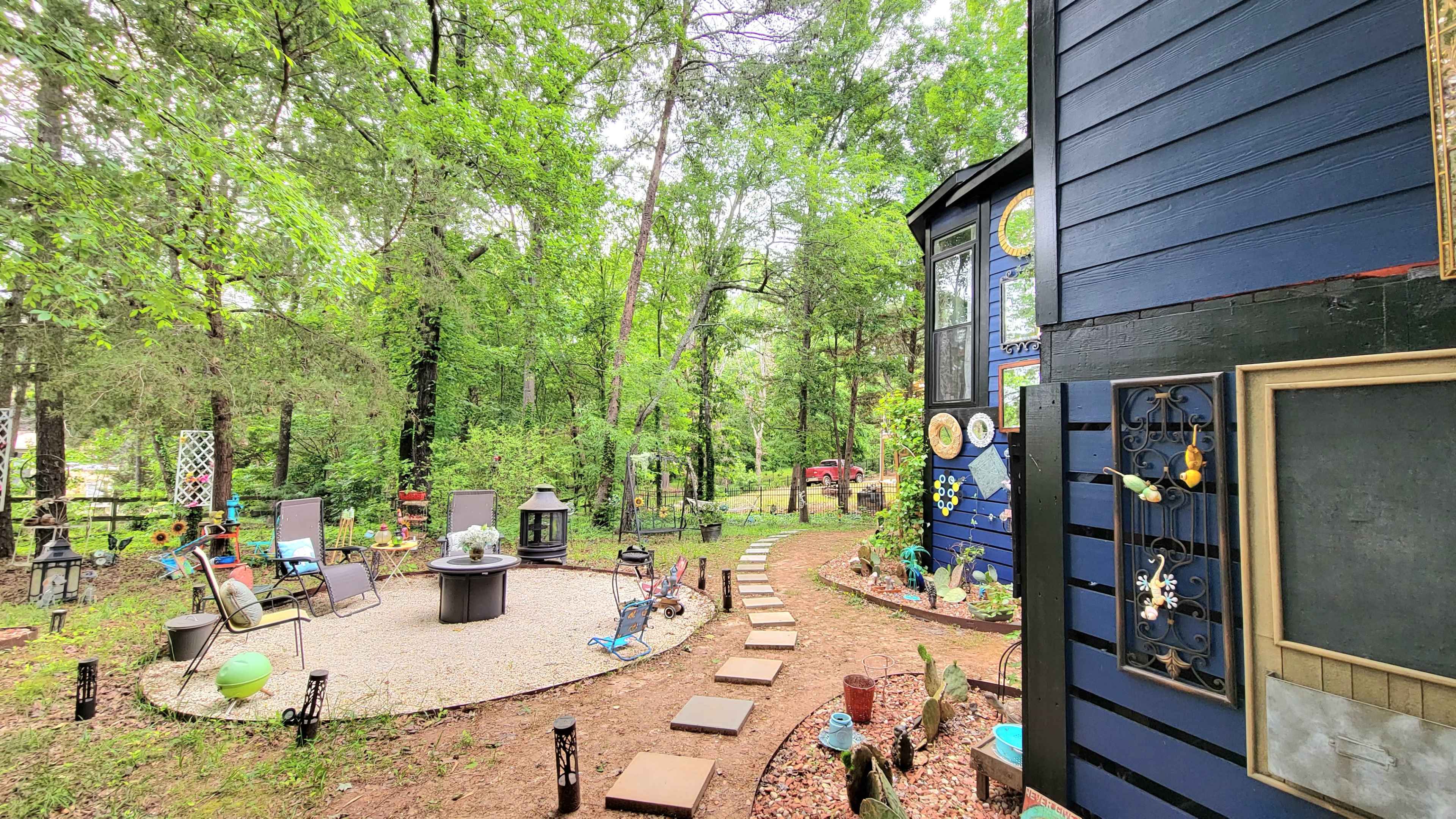 A winding stone path leads through a landscaped garden with various decorations alongside a blue house surrounded by trees.