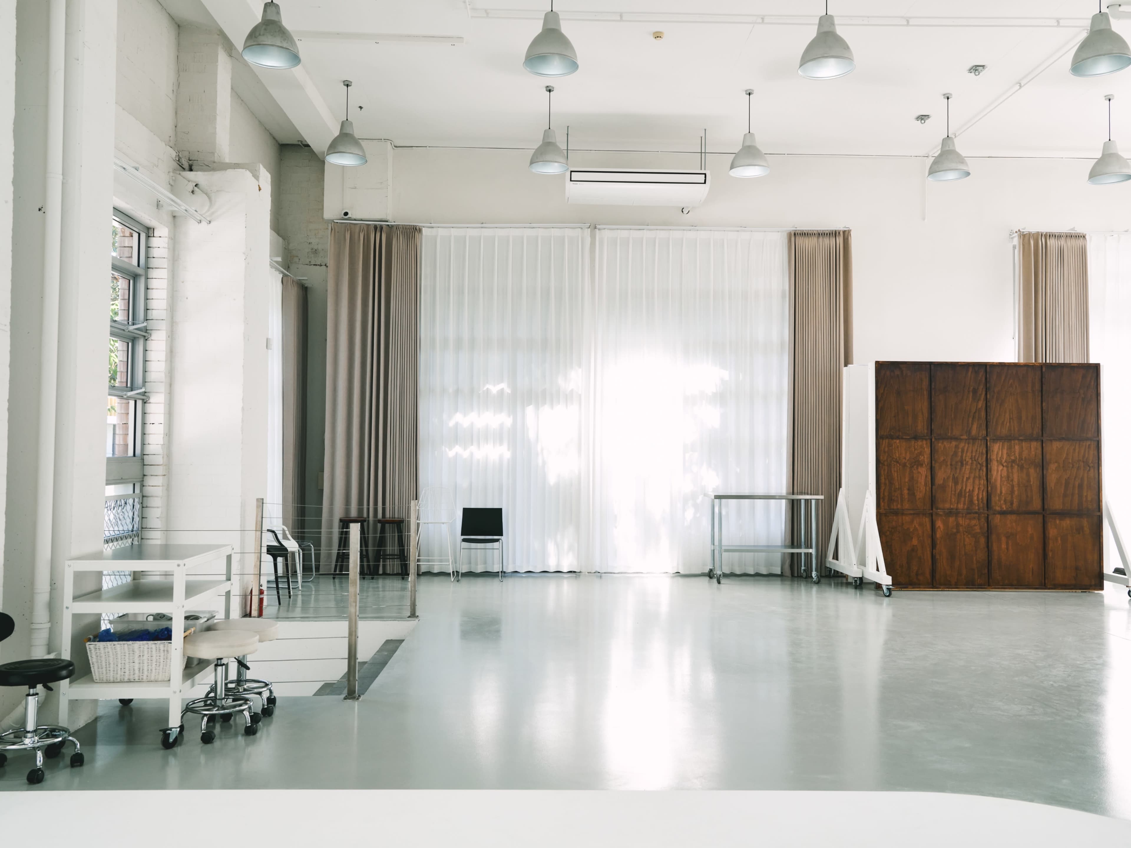The image shows a spacious, well-lit studio with large windows, light curtains, and wooden partitions.