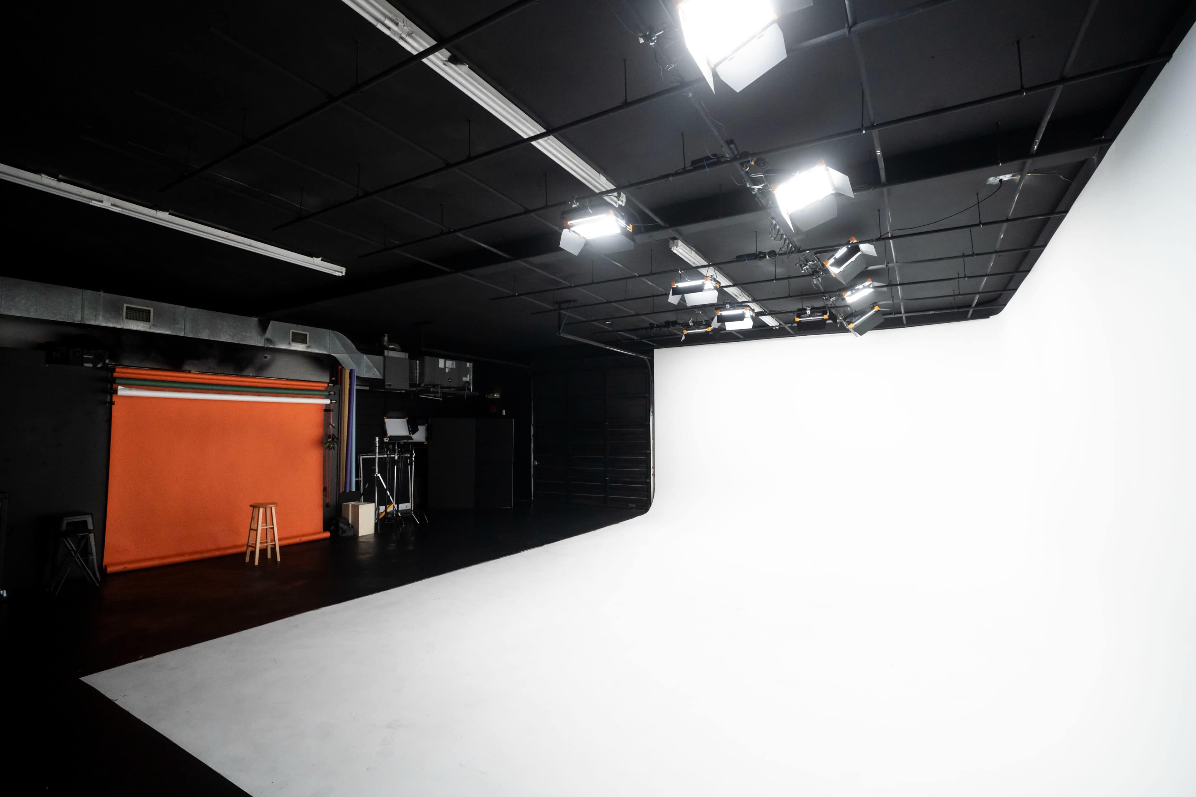 A photography studio features an orange backdrop, a stool, and multiple overhead lights illuminating a white backdrop area.