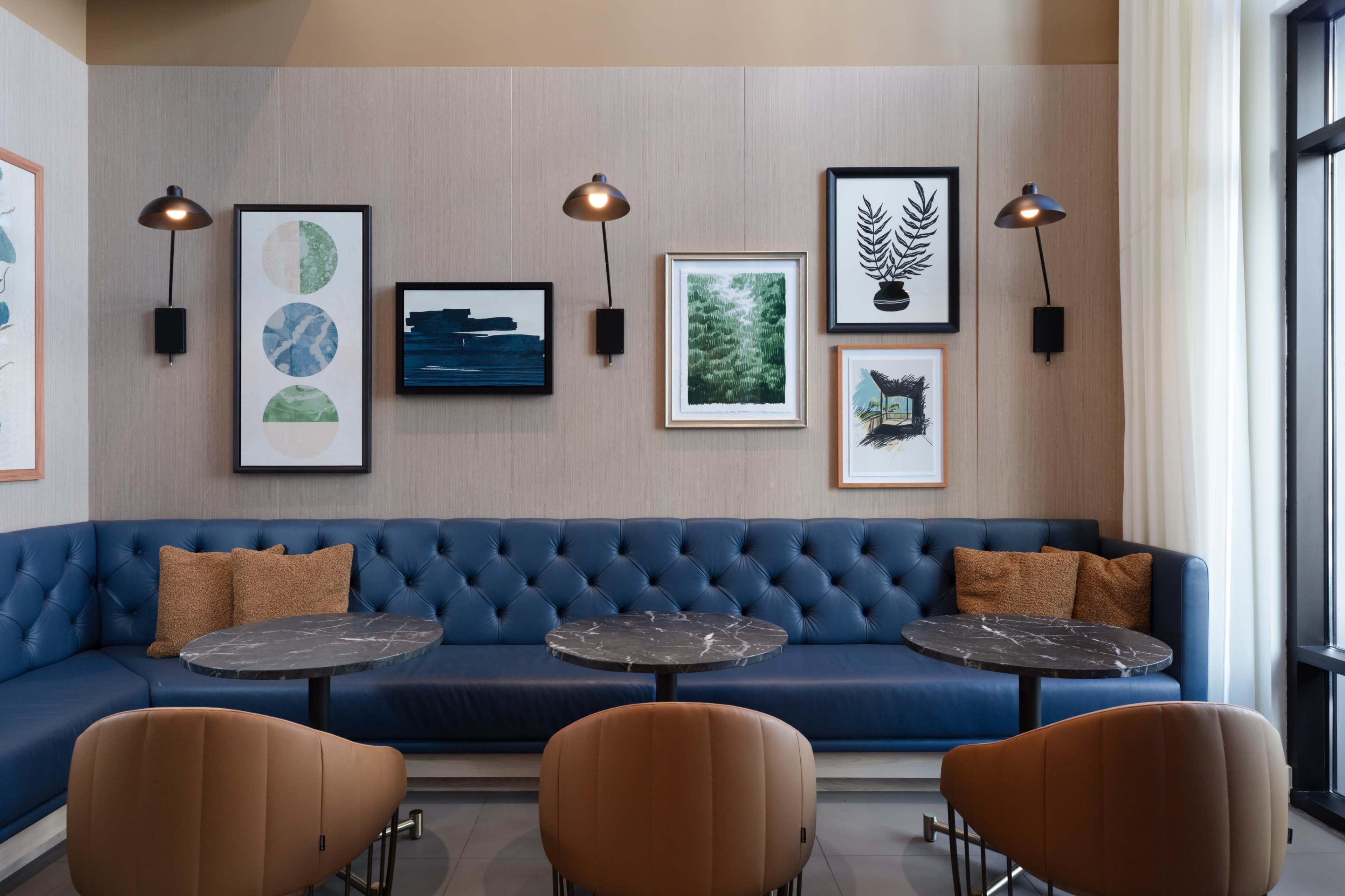 The image shows a modern lounge area with a blue tufted bench, marble-topped tables, and framed artwork on the walls.