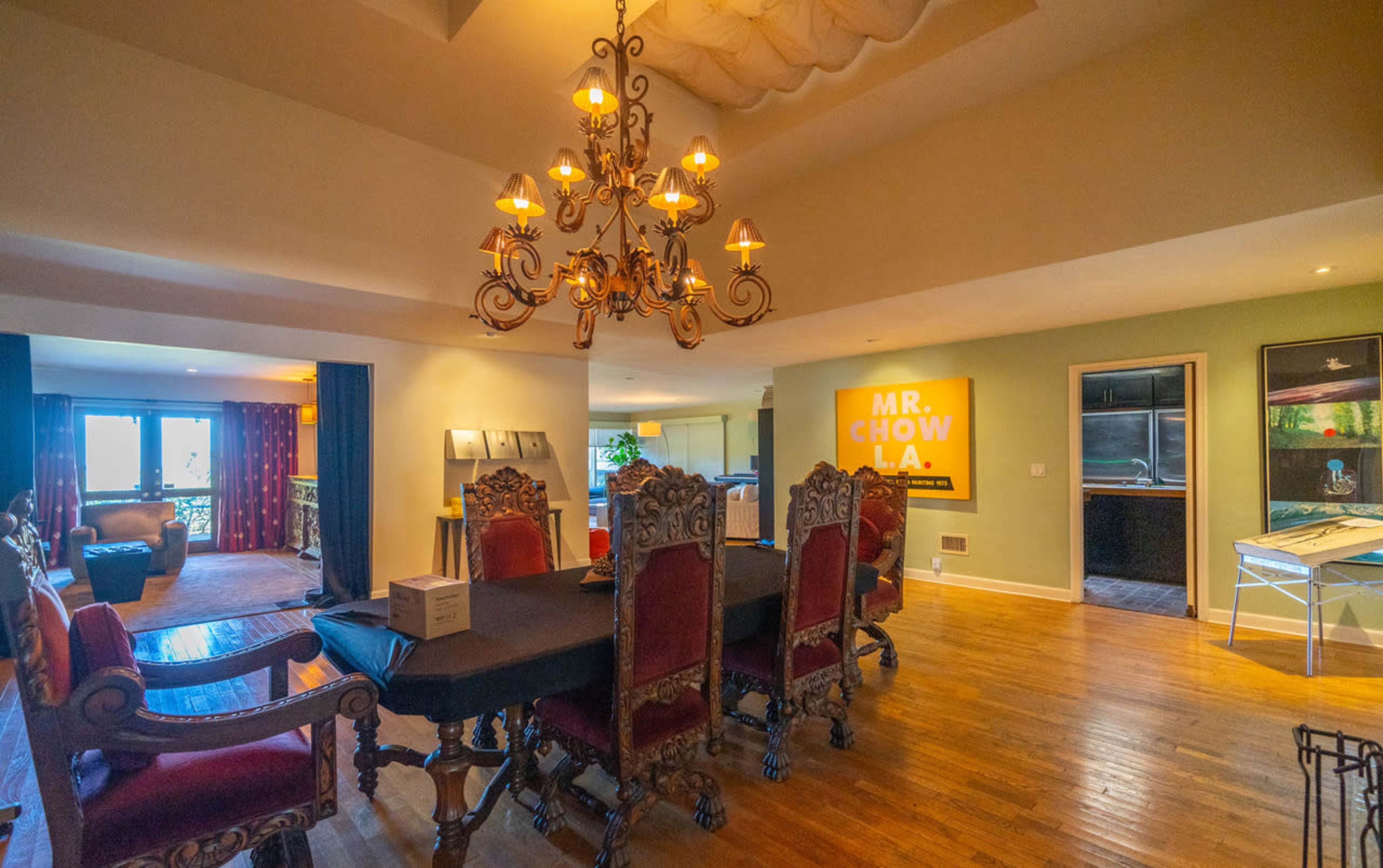 The scene features a dining area with an ornate chandelier above a large table surrounded by intricately designed chairs, leading into a living space with a couch and artwork on the walls.