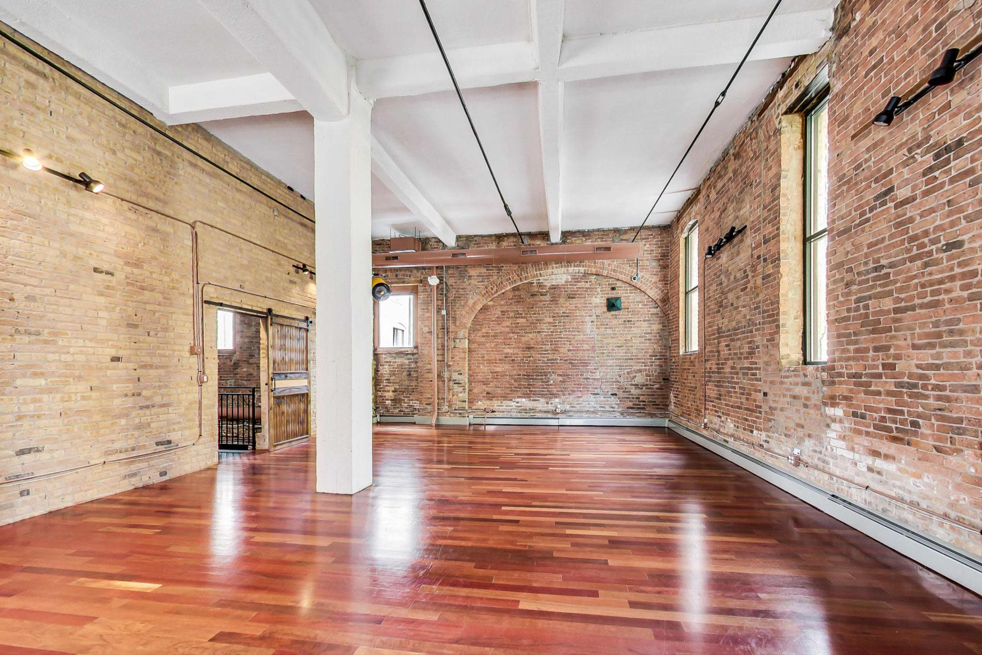 A spacious interior with exposed brick walls and polished wooden flooring, featuring high ceilings and large windows.