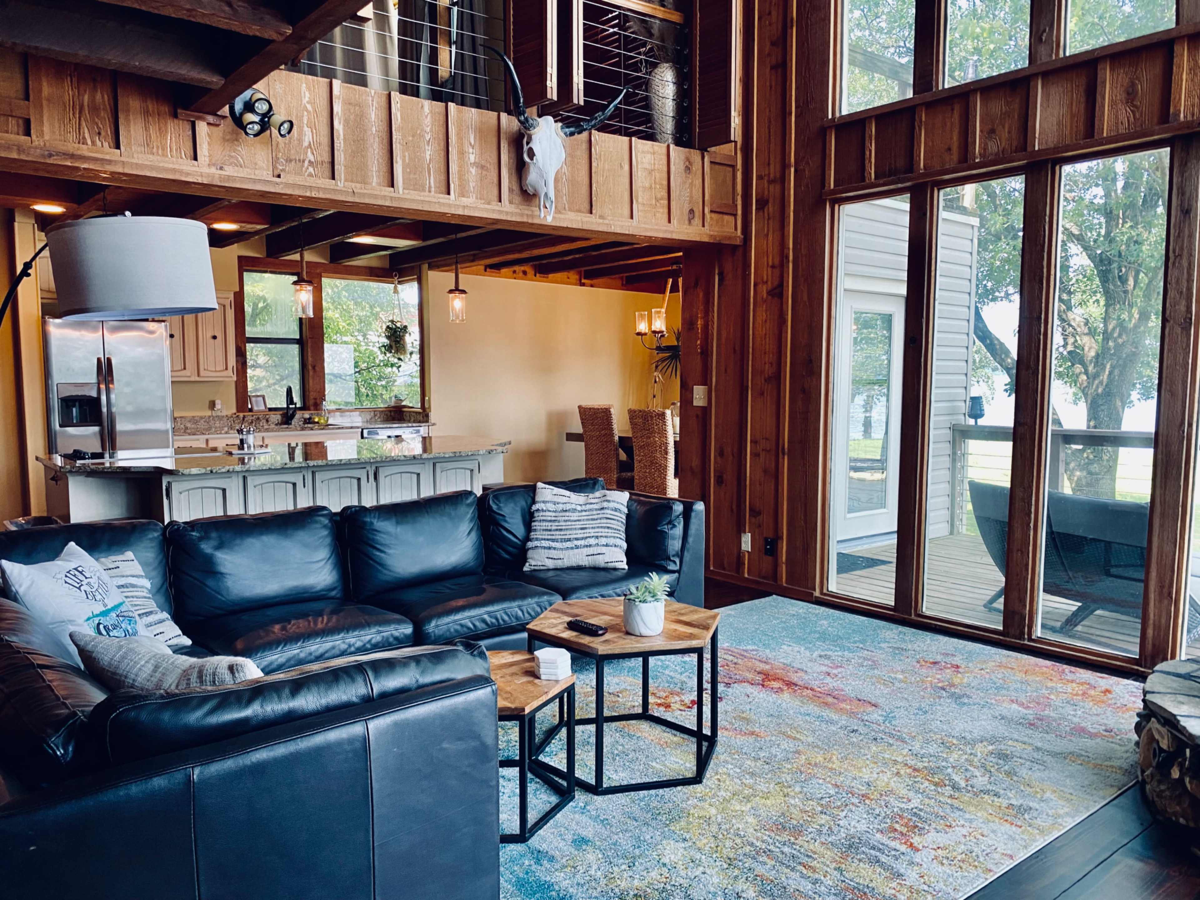 The image shows a spacious living room with a large black sectional sofa, wooden accents, and a view of the outdoors through large windows.