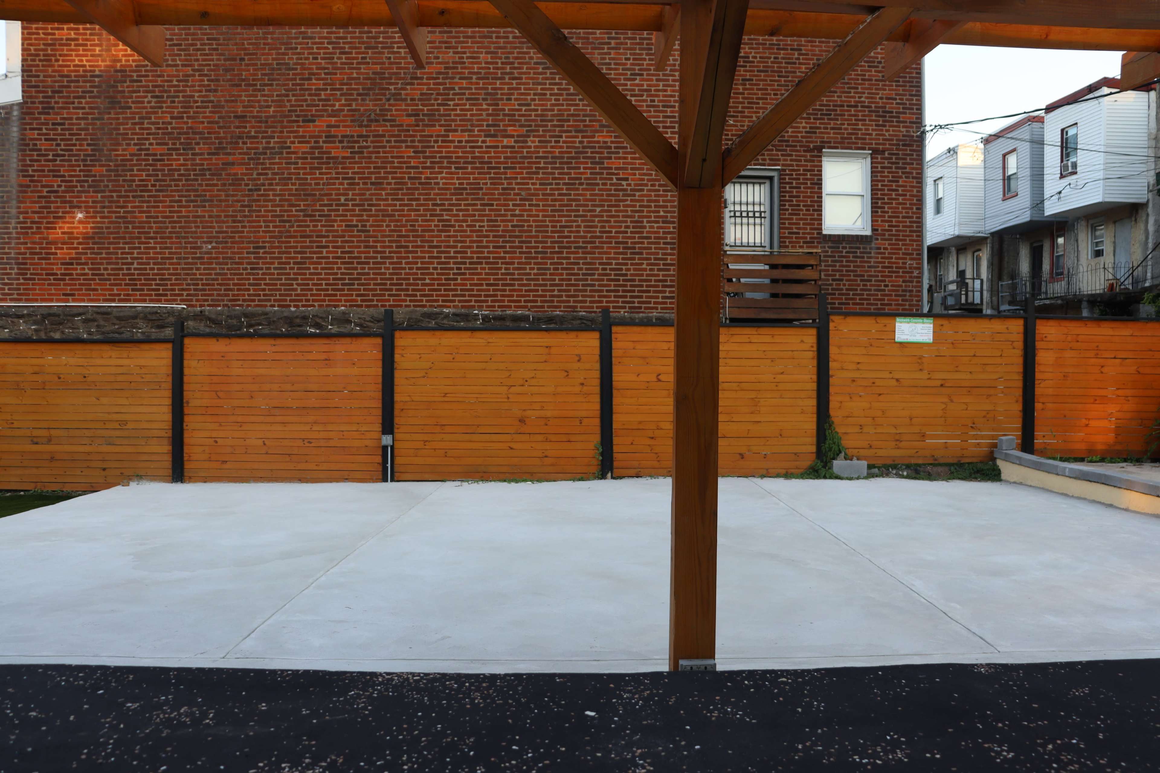 The image shows a concrete area with a wooden support structure, bordered by wooden fences and a brick building in the background.