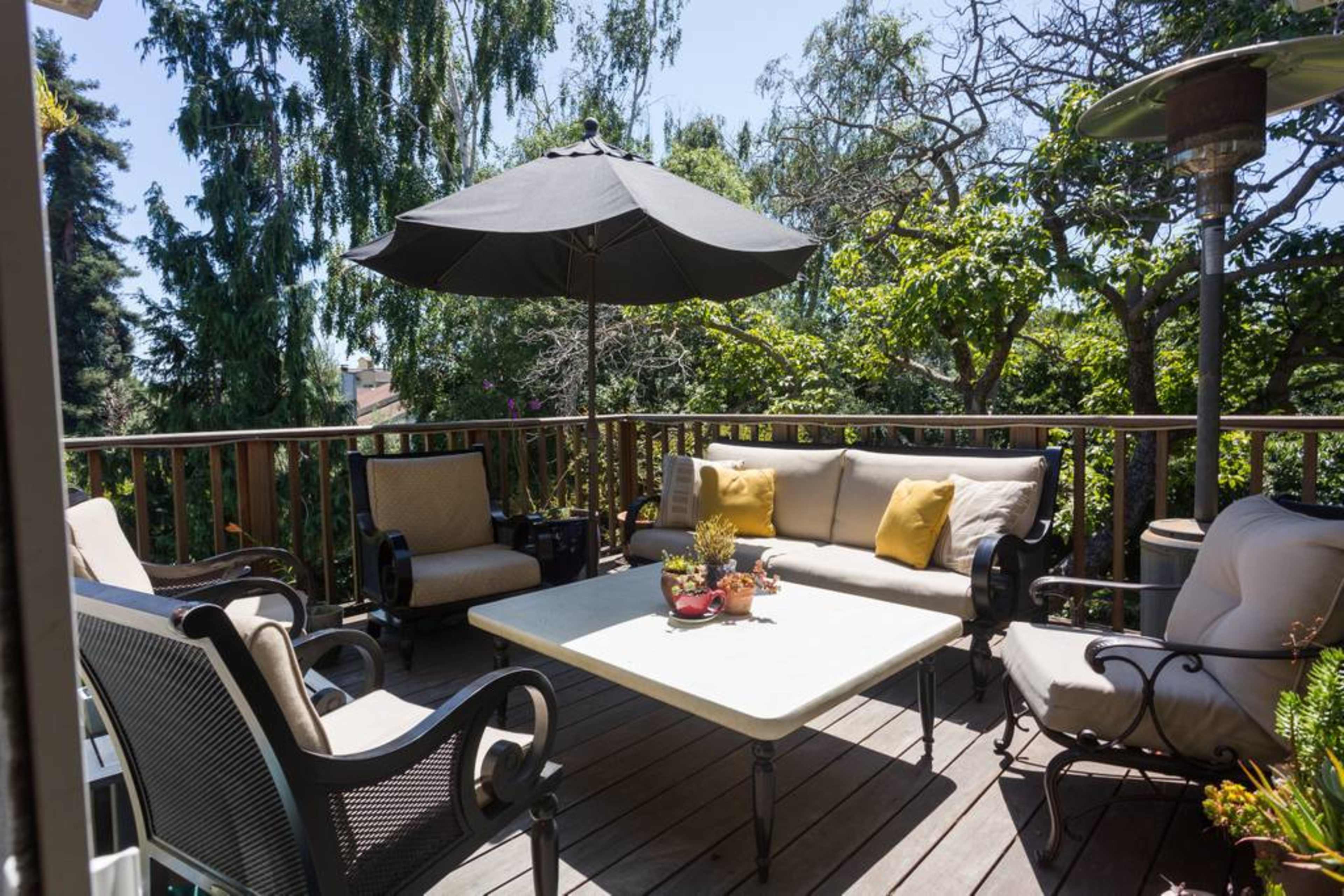 A patio area features a table and several chairs beneath a large umbrella, surrounded by greenery.