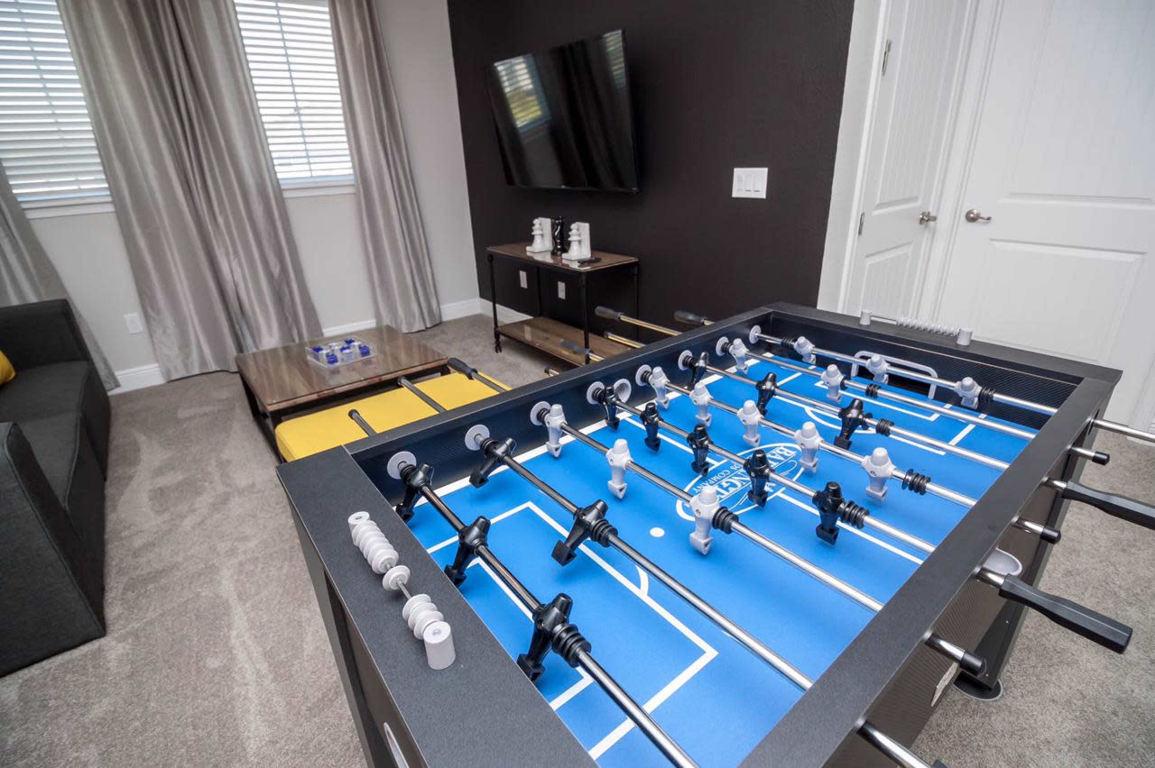 A foosball table in a game room with a gray sofa, a television mounted on the wall, and a coffee table nearby.