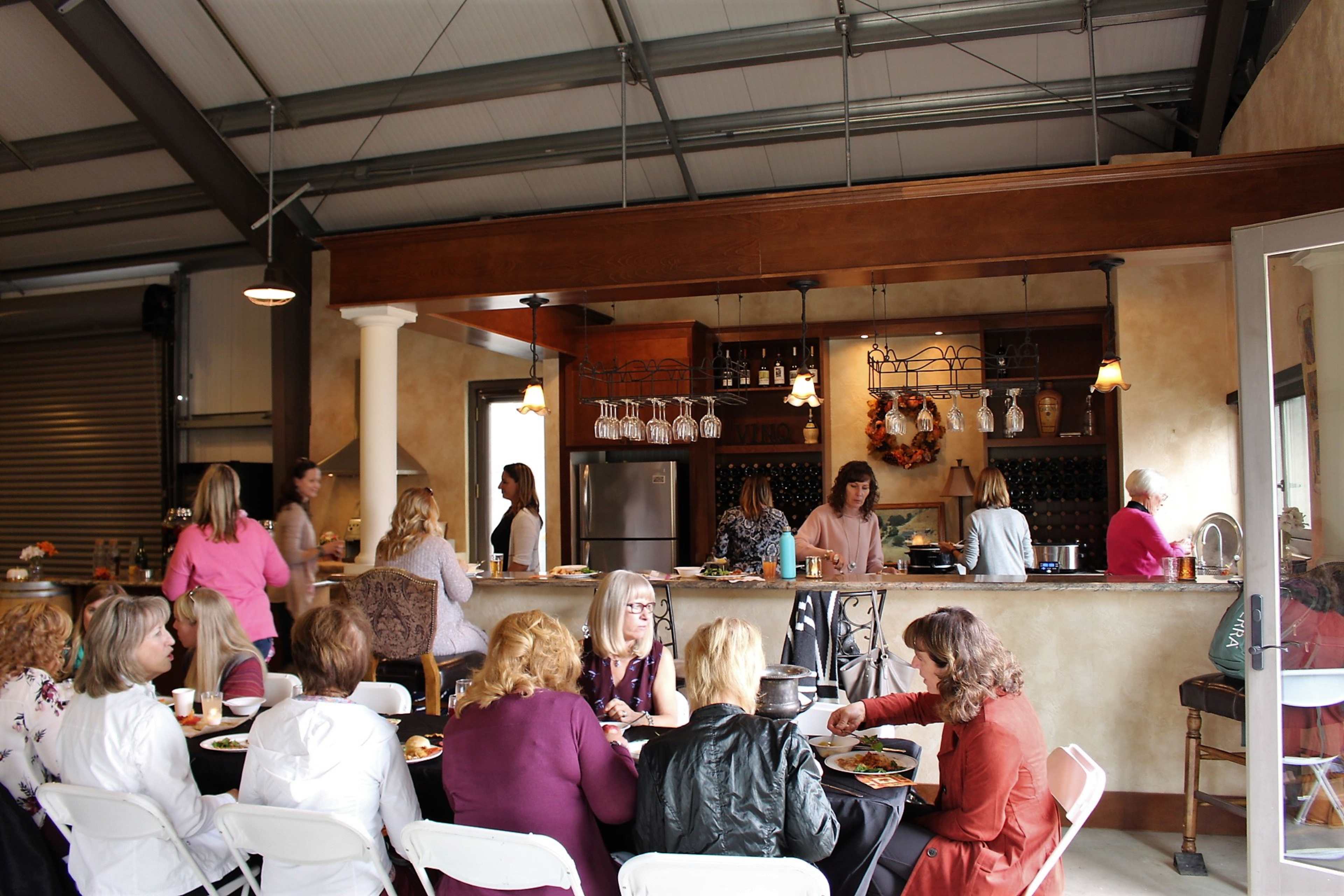A group of women sits at tables with food in a spacious dining area, while others prepare food and serve drinks at the bar in the background.