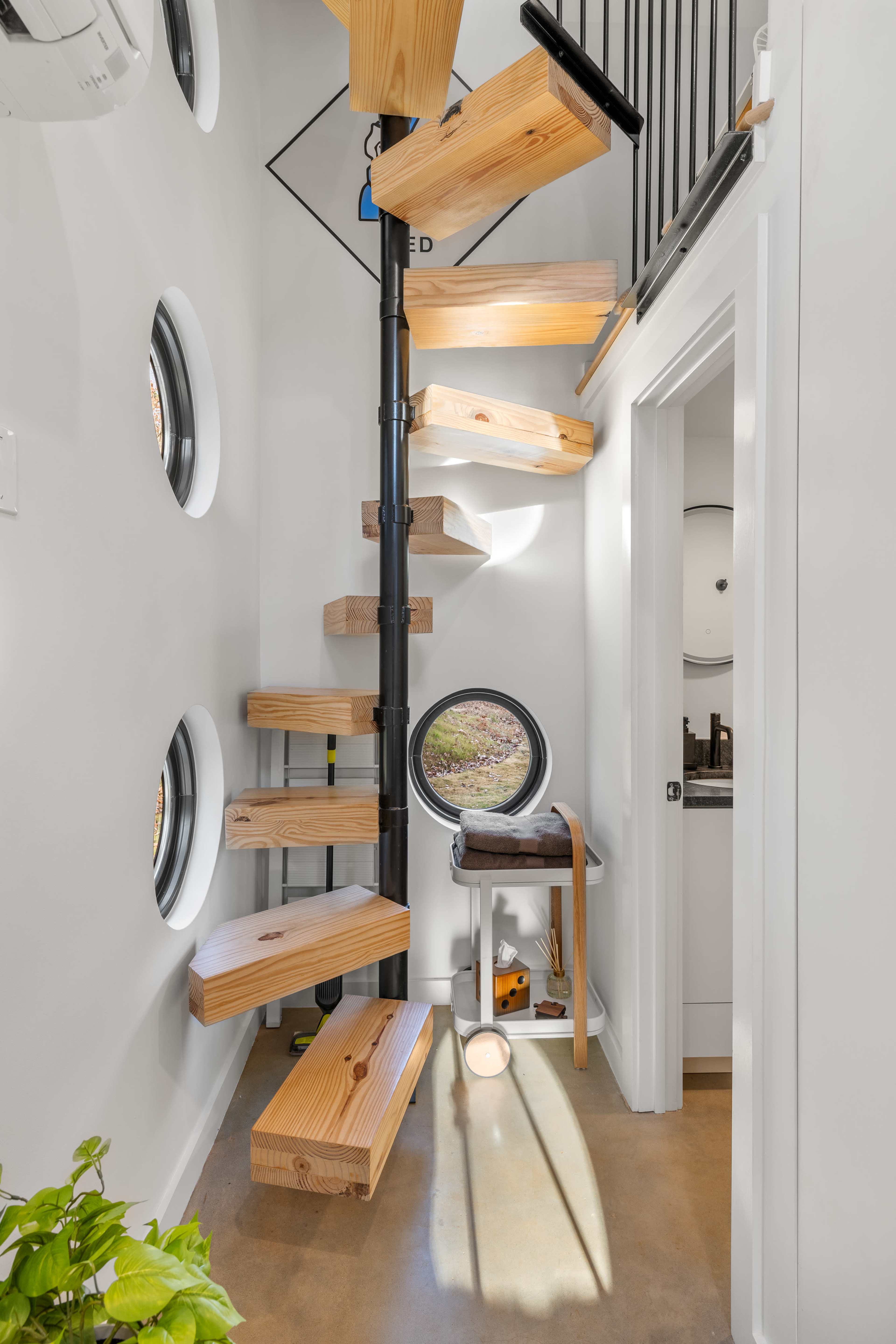 The image shows a modern spiral staircase made of wooden steps, leading upwards in a bright, white-walled interior with circular windows.