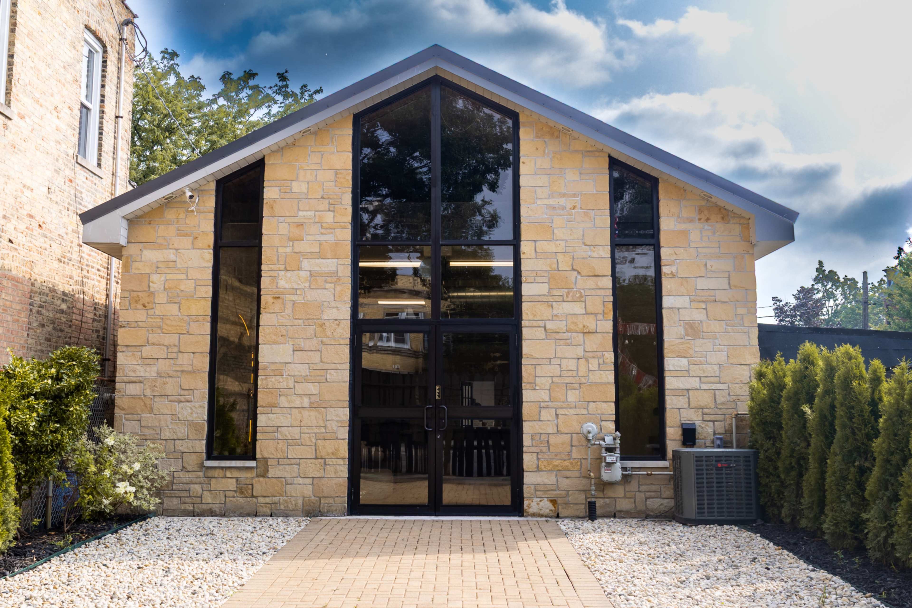 The image shows a modern building with a stone facade and large glass windows, situated in a landscaped setting with gravel pathways.