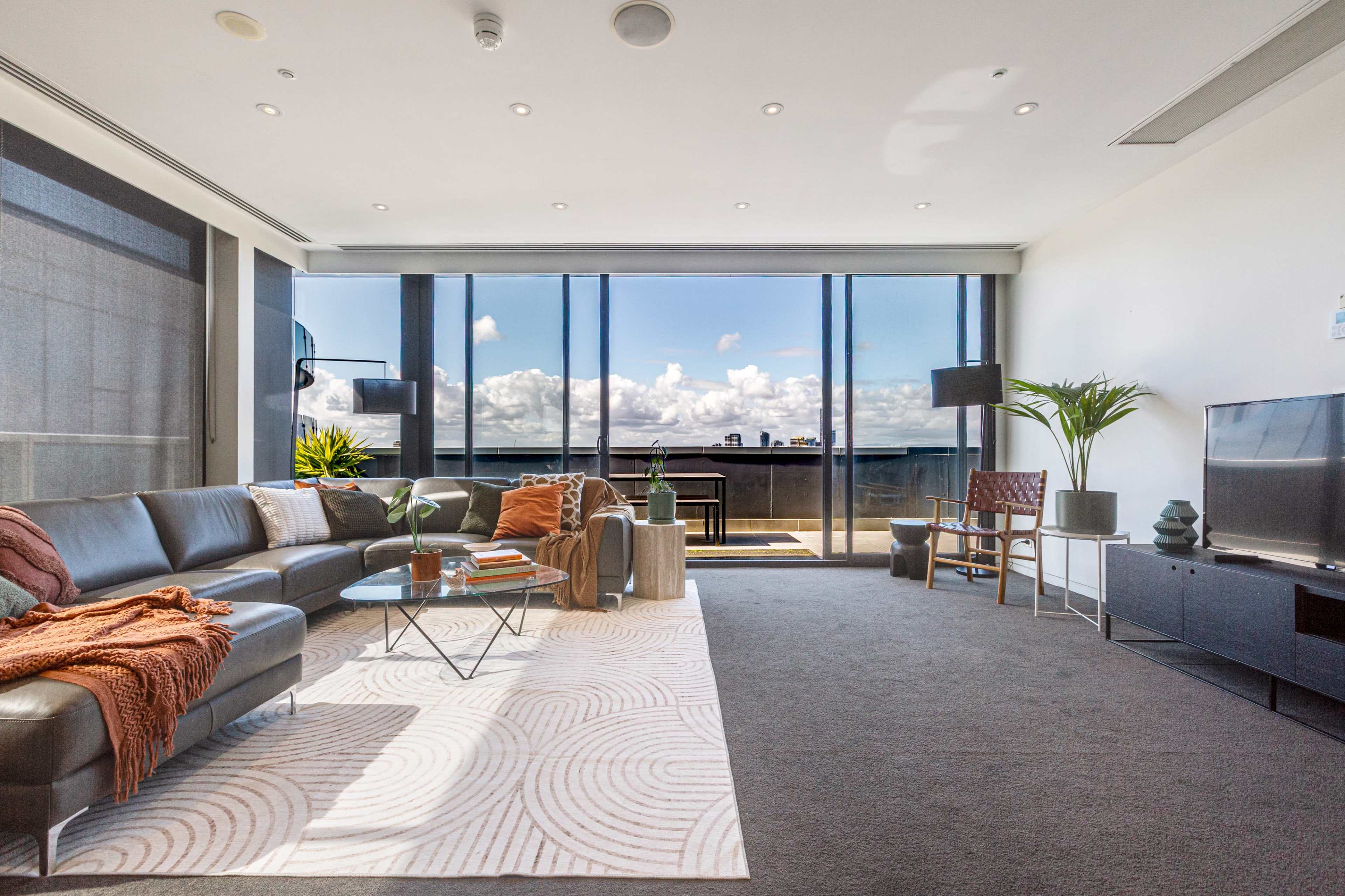 The image depicts a modern living room with a large sectional sofa, a coffee table, and a view of a balcony overlooking a bright sky with clouds.