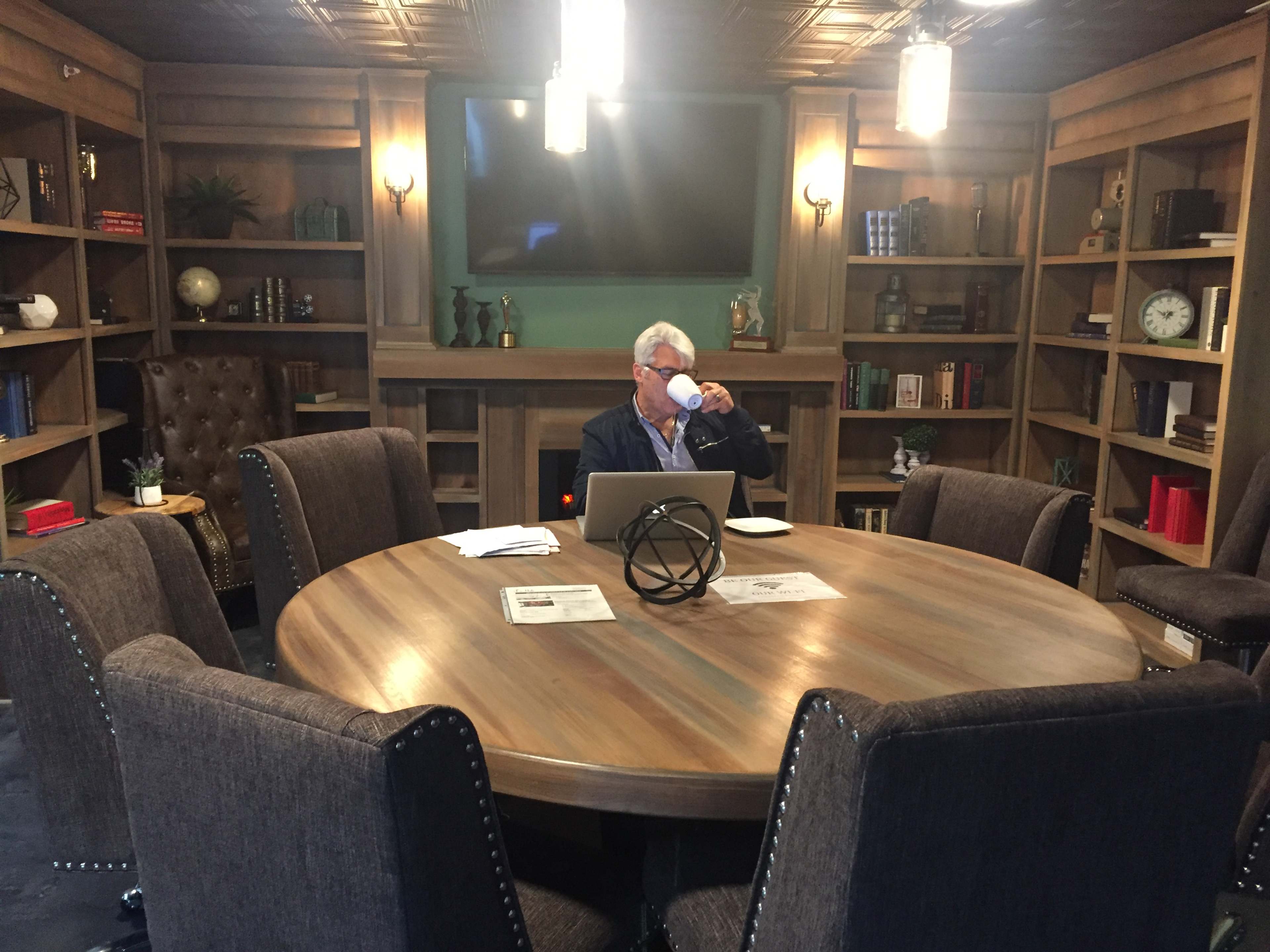 A man sits at a round wooden table in a well-furnished meeting room, sipping from a mug while working on a laptop.