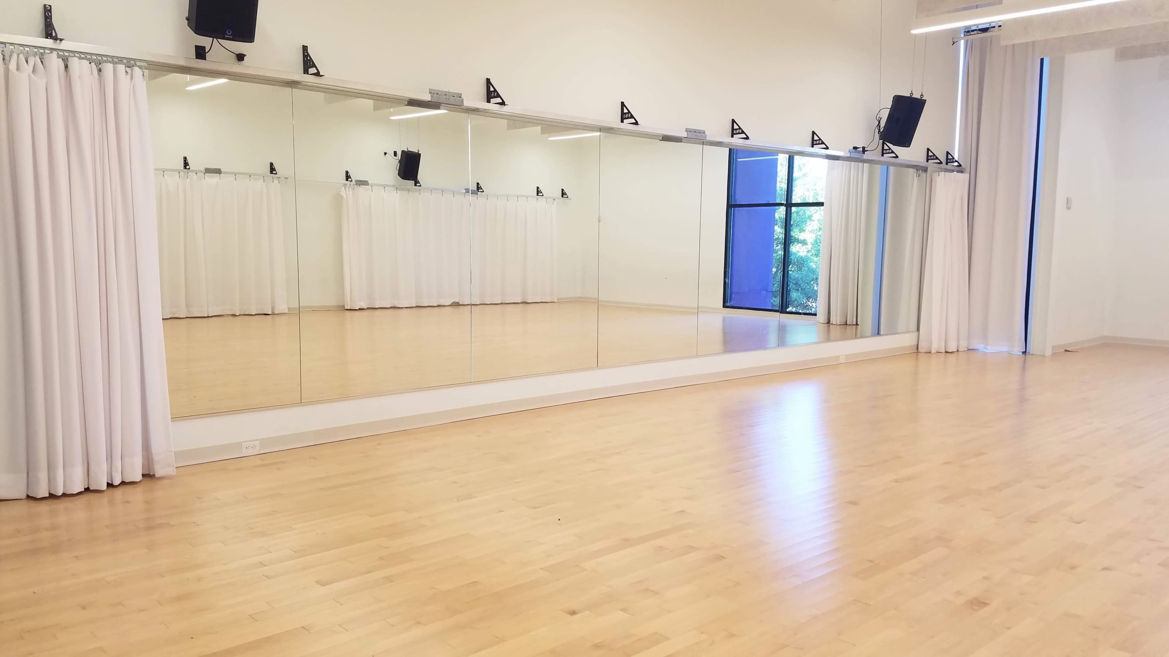 The image shows a dance studio with wooden floors, large mirrors, and white curtains along one wall.