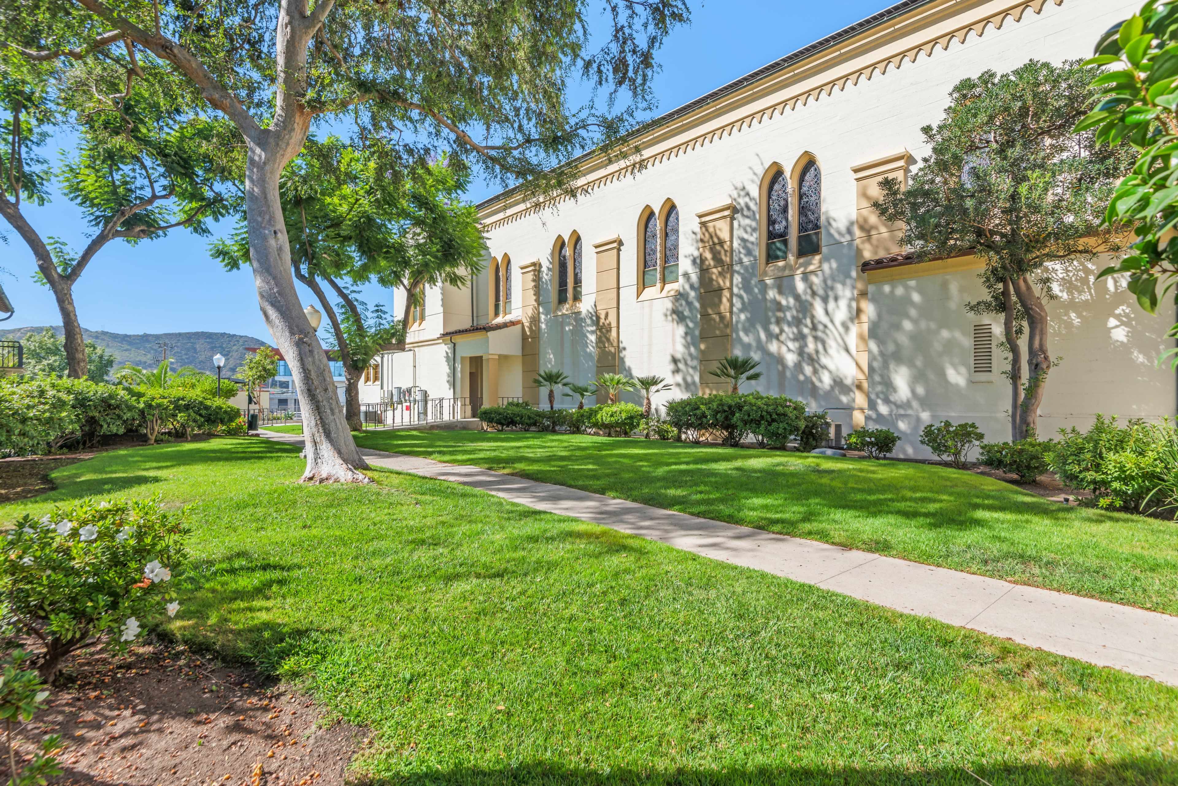 The image shows a landscaped area with a pathway leading to a white building, surrounded by greenery and trees under a clear blue sky.