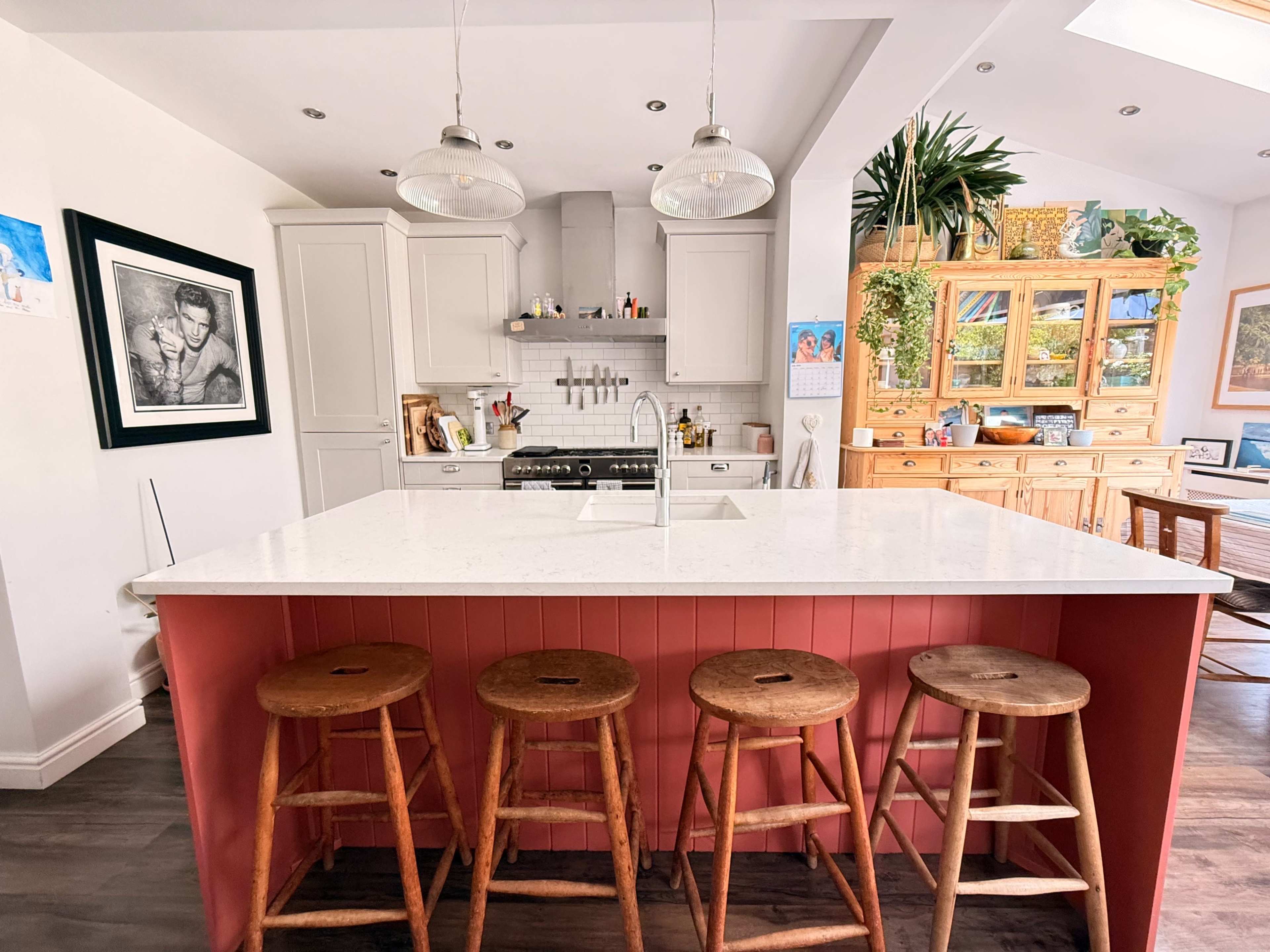 The image shows a modern kitchen with a white countertop island, four wooden bar stools, and a mix of cabinetry and shelves featuring various decorations and plants.