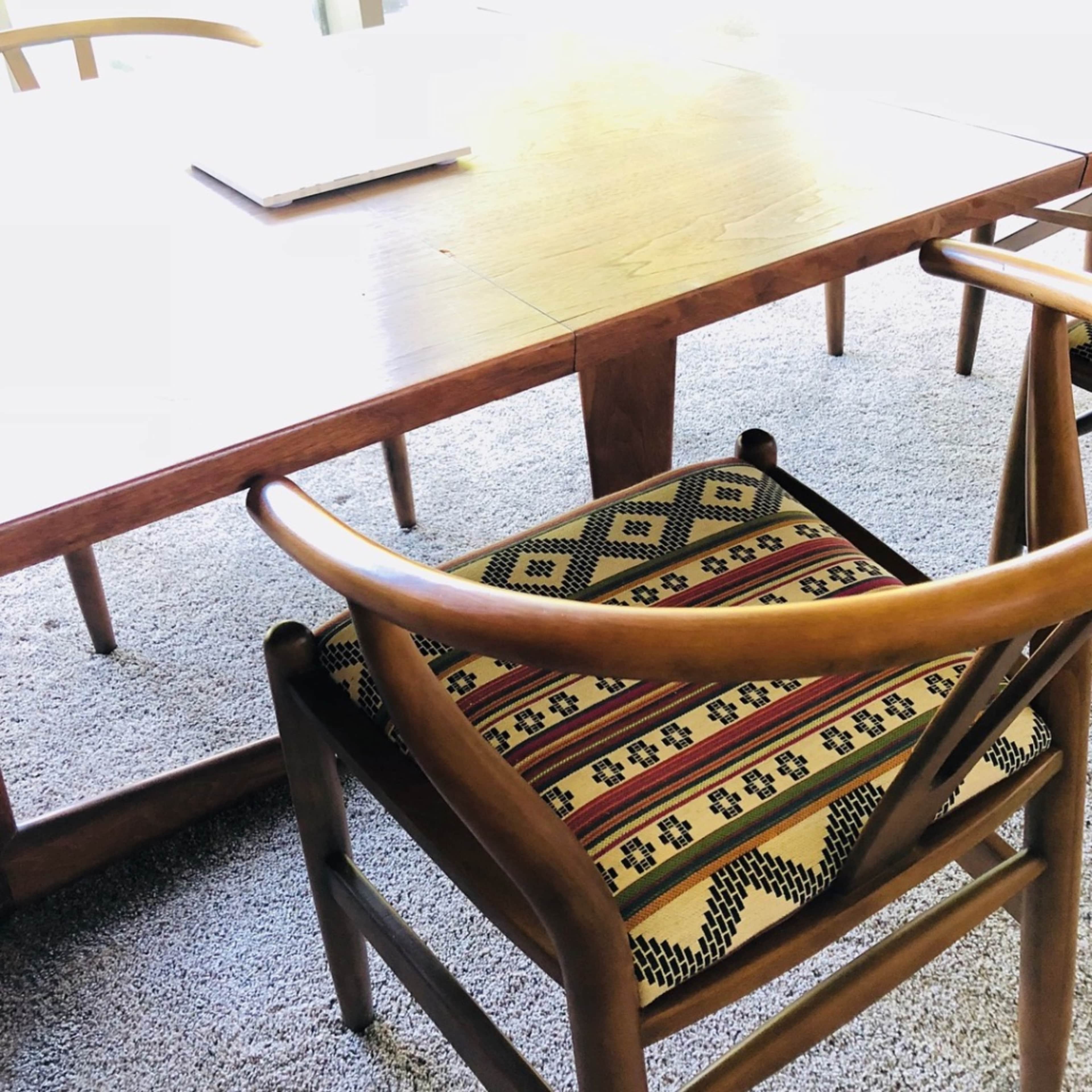 A wooden table with a laptop sits beside a chair featuring a patterned cushion on a carpeted floor.