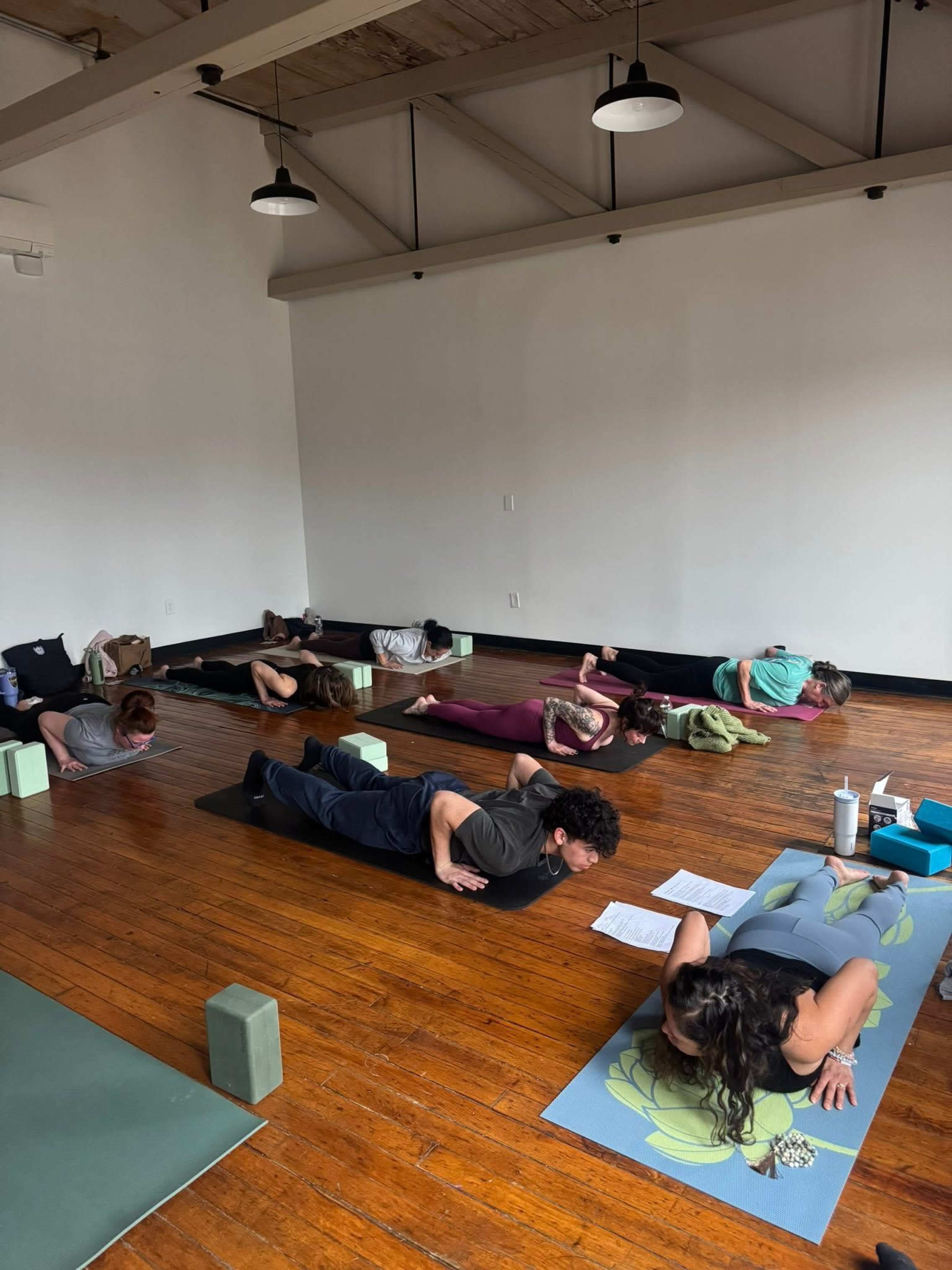 A group of people is practicing yoga on mats in a spacious, well-lit studio with wooden floors.