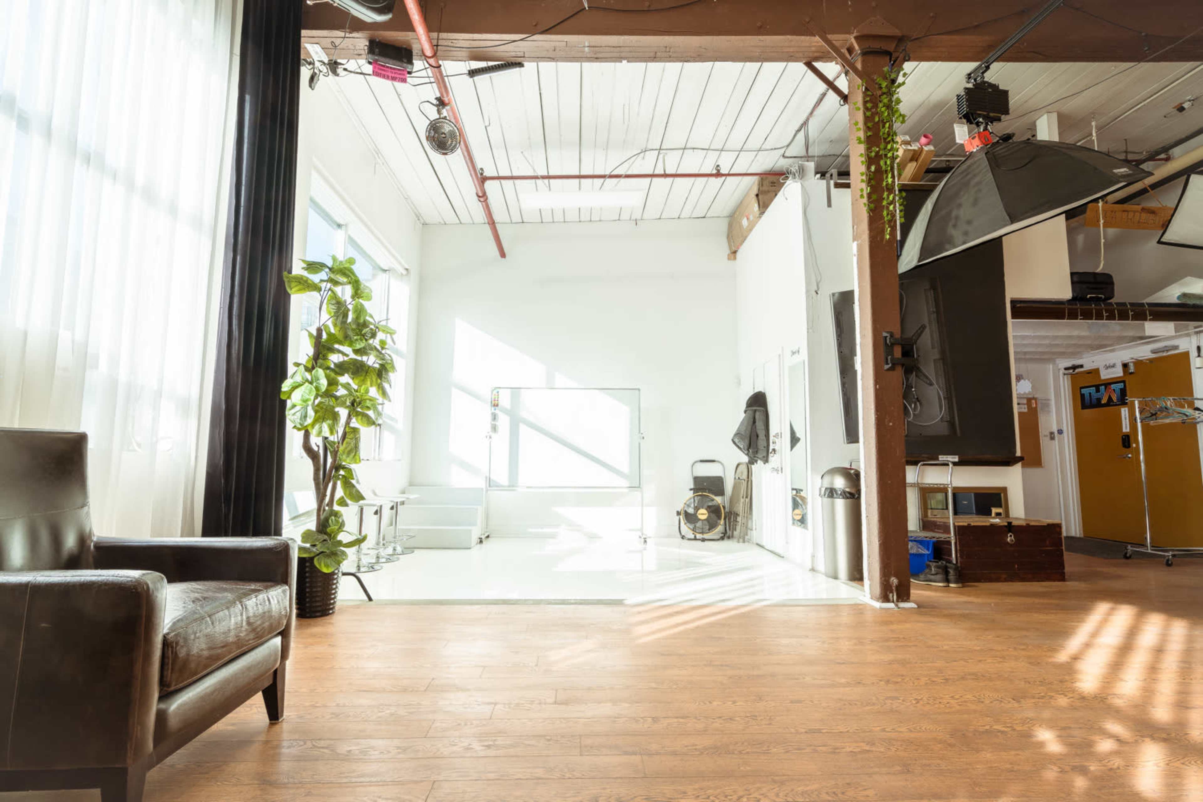 A bright, spacious room with large windows, wood flooring, a potted plant, and some furniture in one corner.