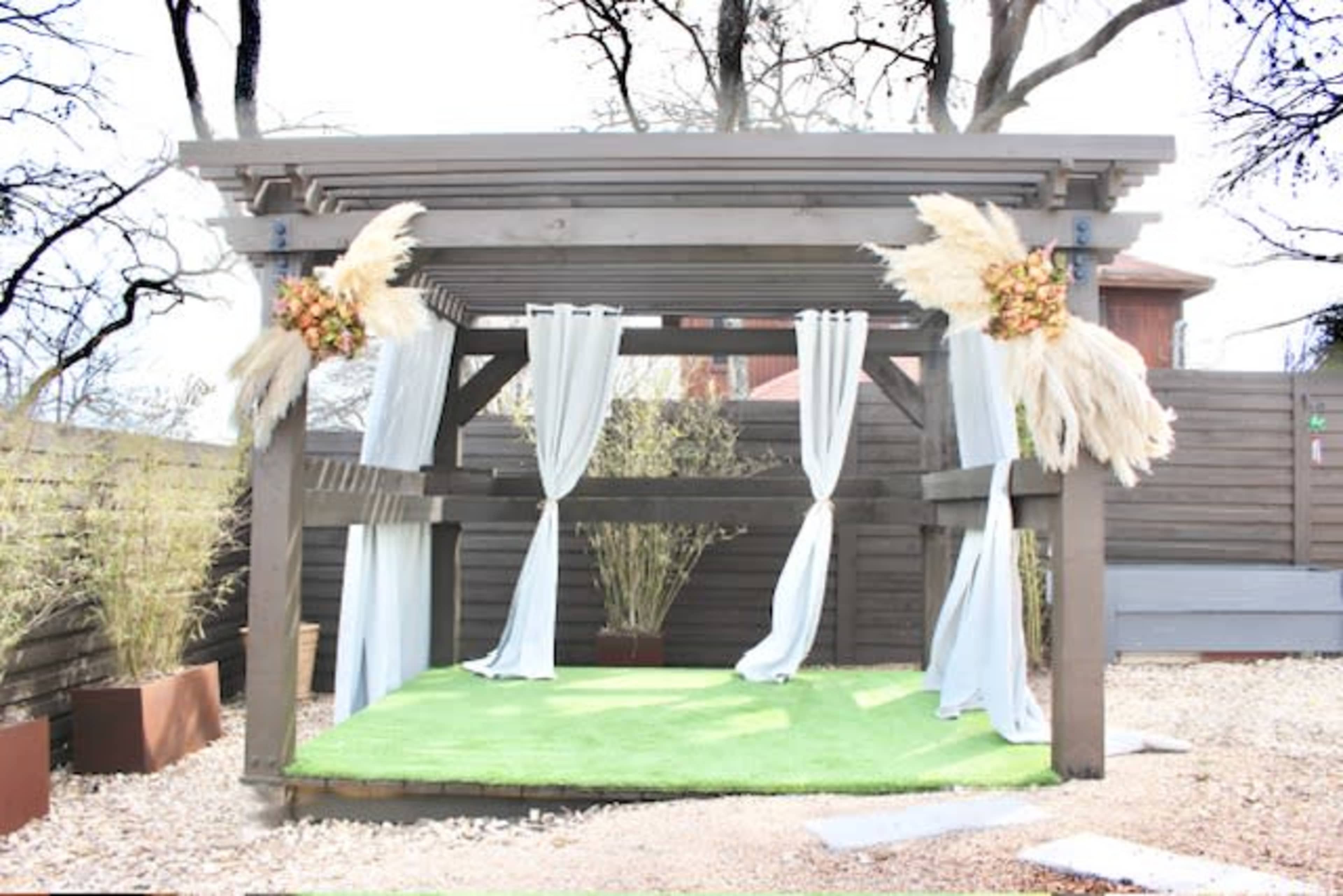 A wooden gazebo with white curtains and decorative plants is set on a grassy area surrounded by a landscaped yard.