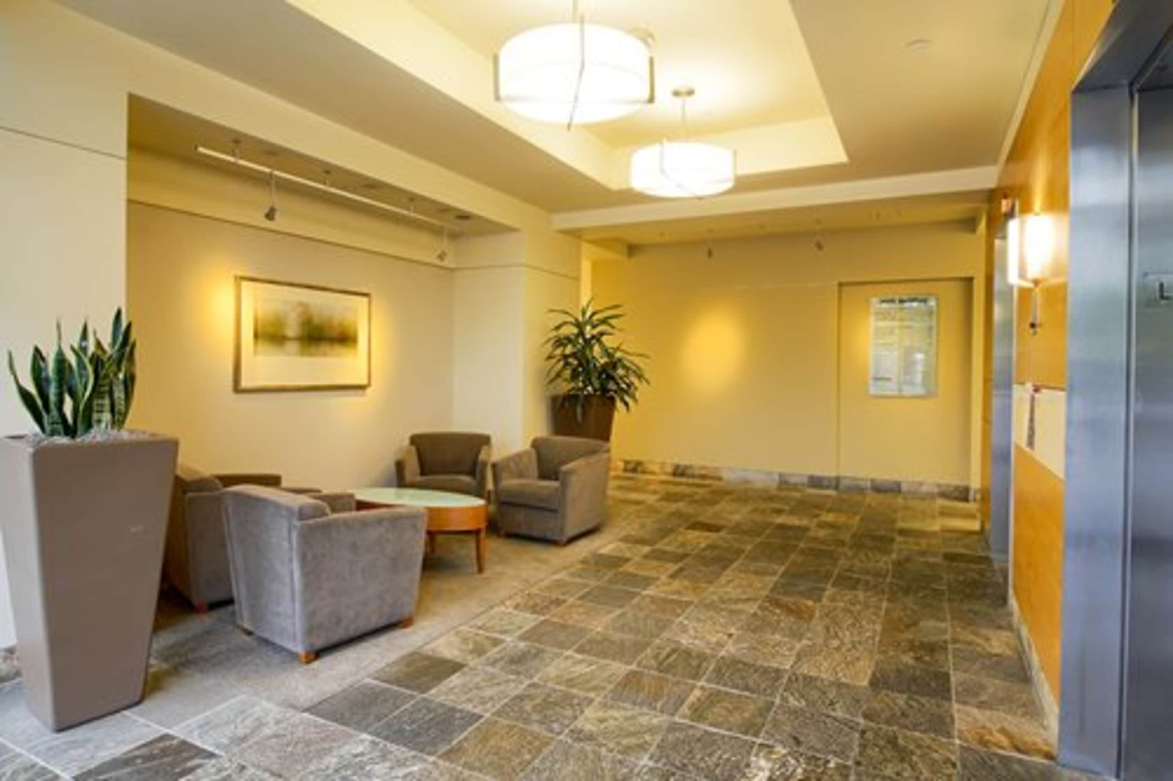 A spacious lobby area with two gray chairs, a wooden coffee table, large potted plants, and an elevator door in a well-lit space.
