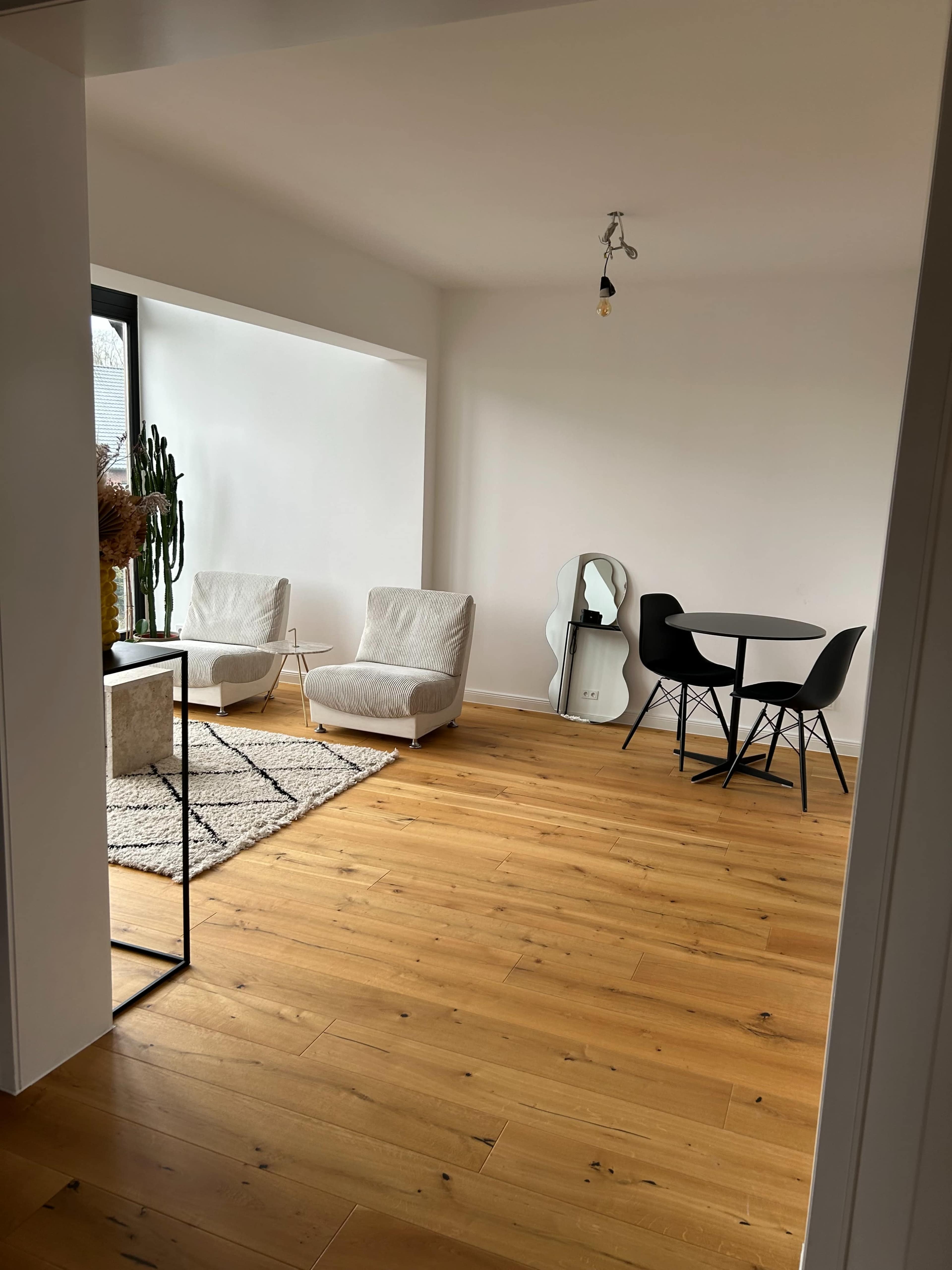 The image shows a bright, minimalistic living space with light wooden flooring, two white chairs, a round black dining table, and a large mirror reflecting the room.