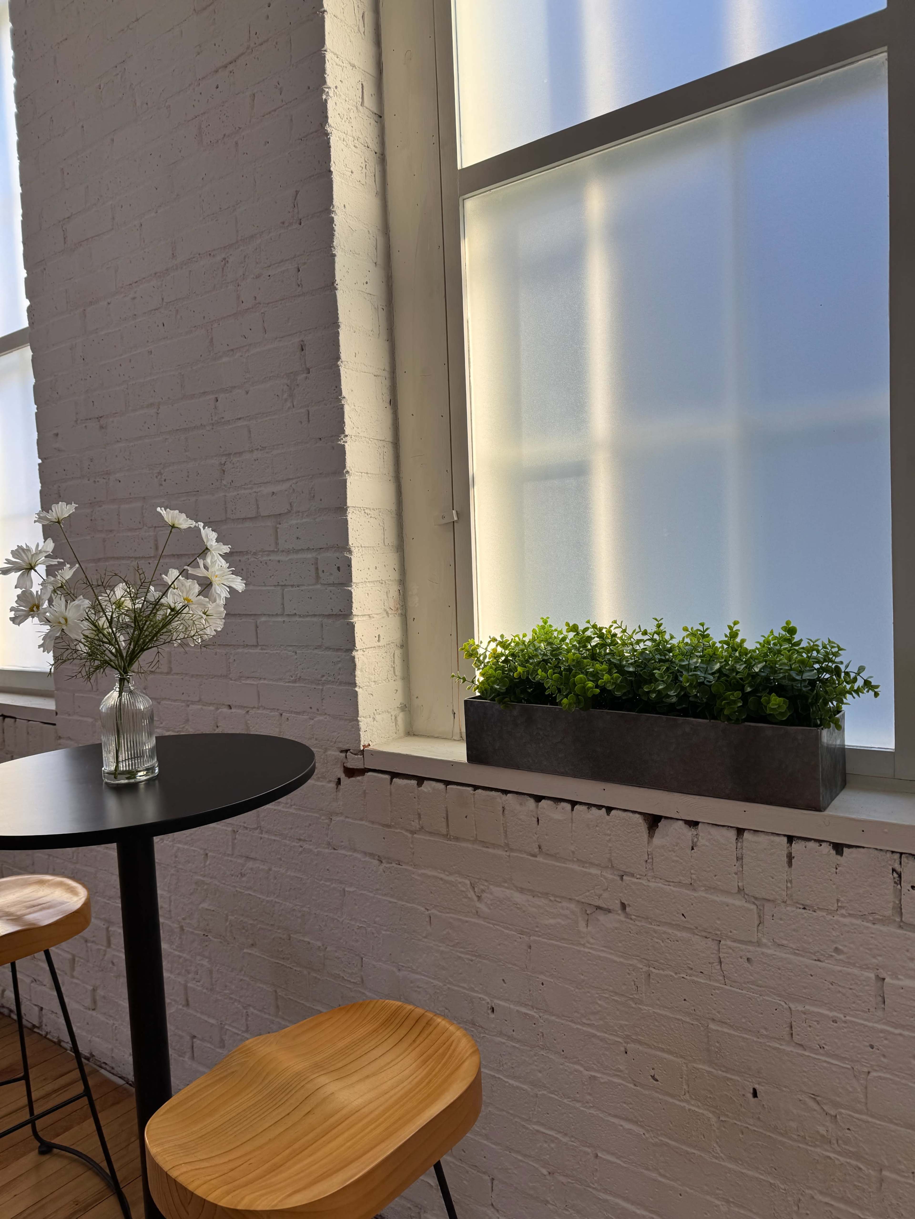 A small black table with two wooden chairs sits next to a window featuring a planter with green plants and a vase of white flowers on the tabletop.