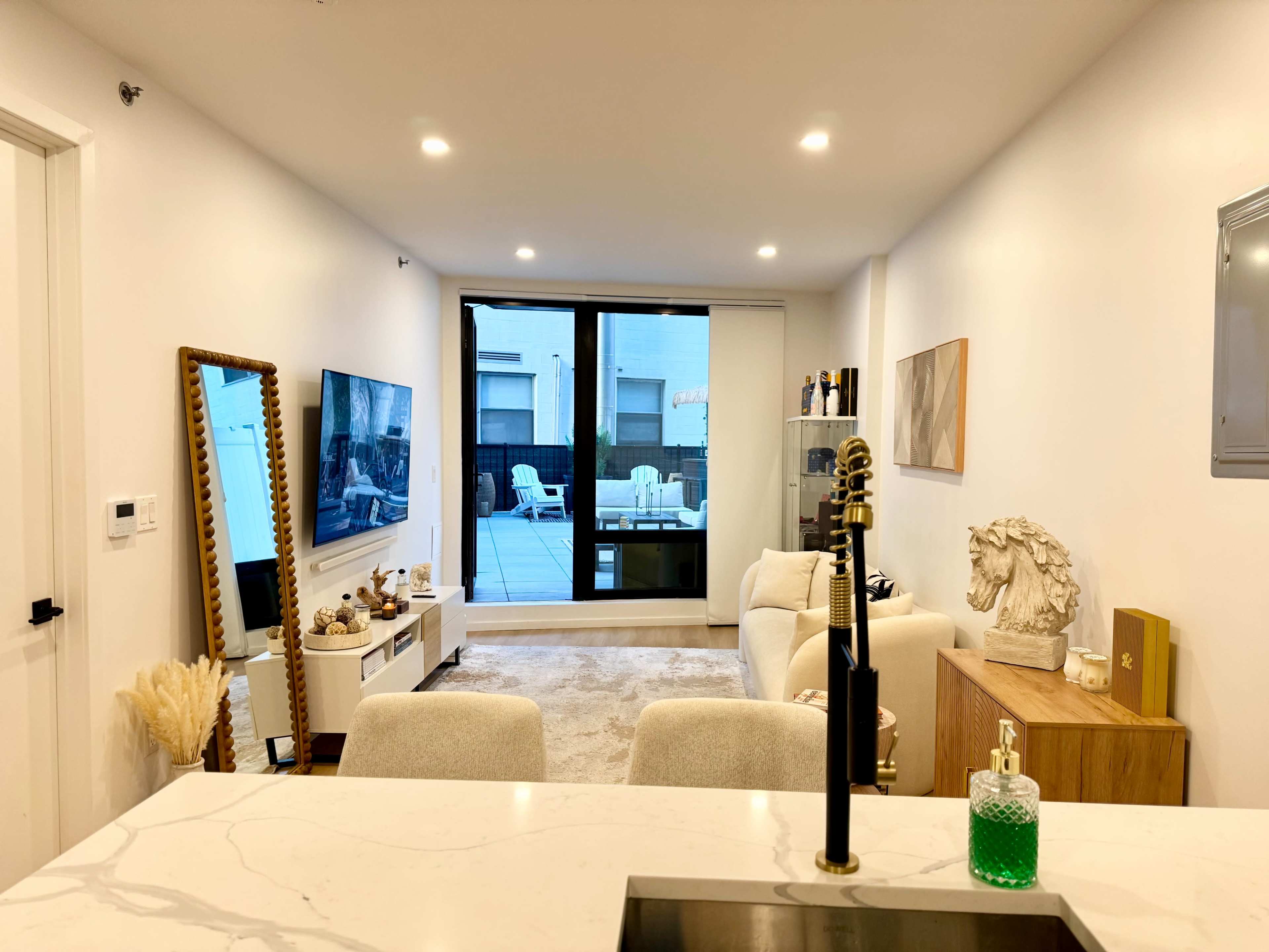 A modern living room features a large window overlooking a patio, with a white sofa, television, and decorative items arranged on shelves.