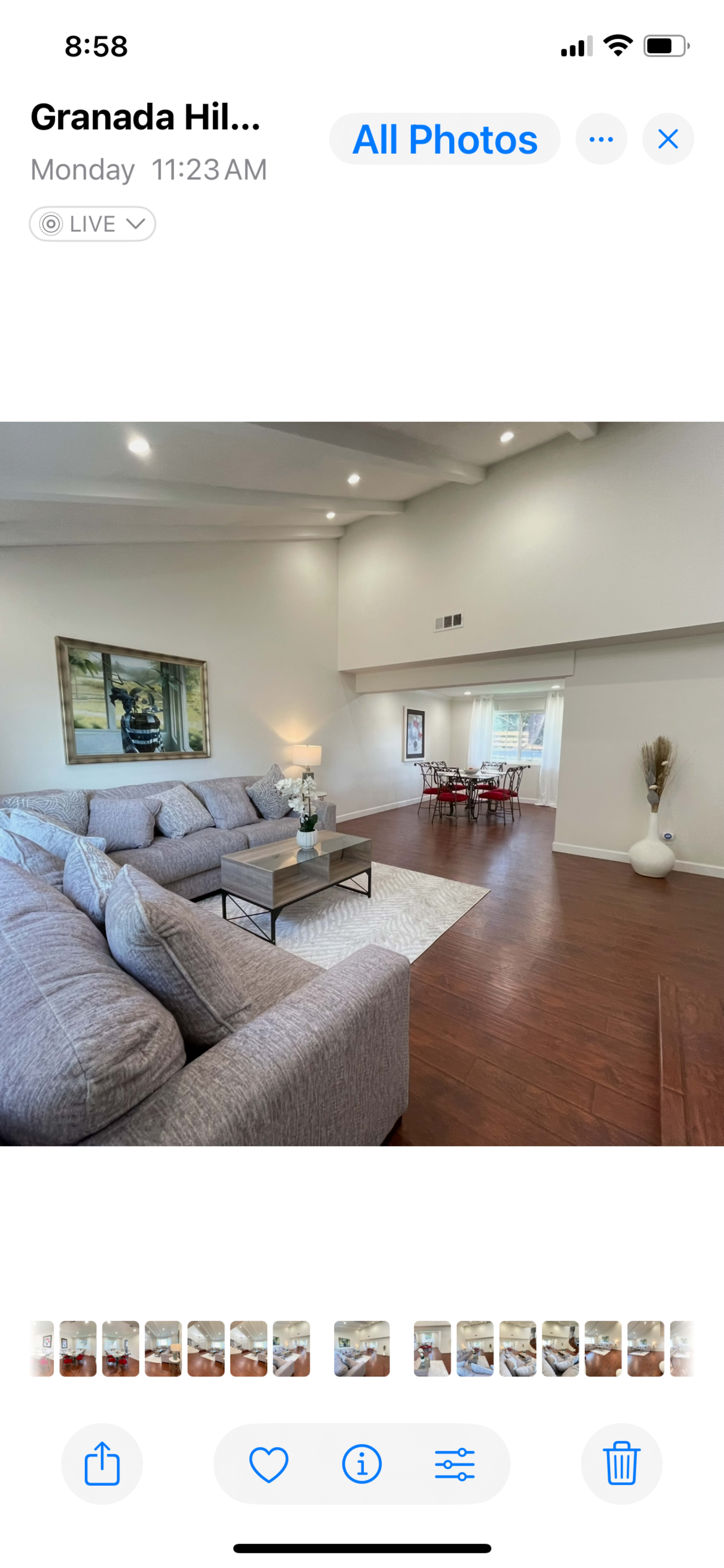 The image shows a spacious living room area featuring a large gray sectional sofa, wooden flooring, artwork on the wall, and a dining area visible in the background.