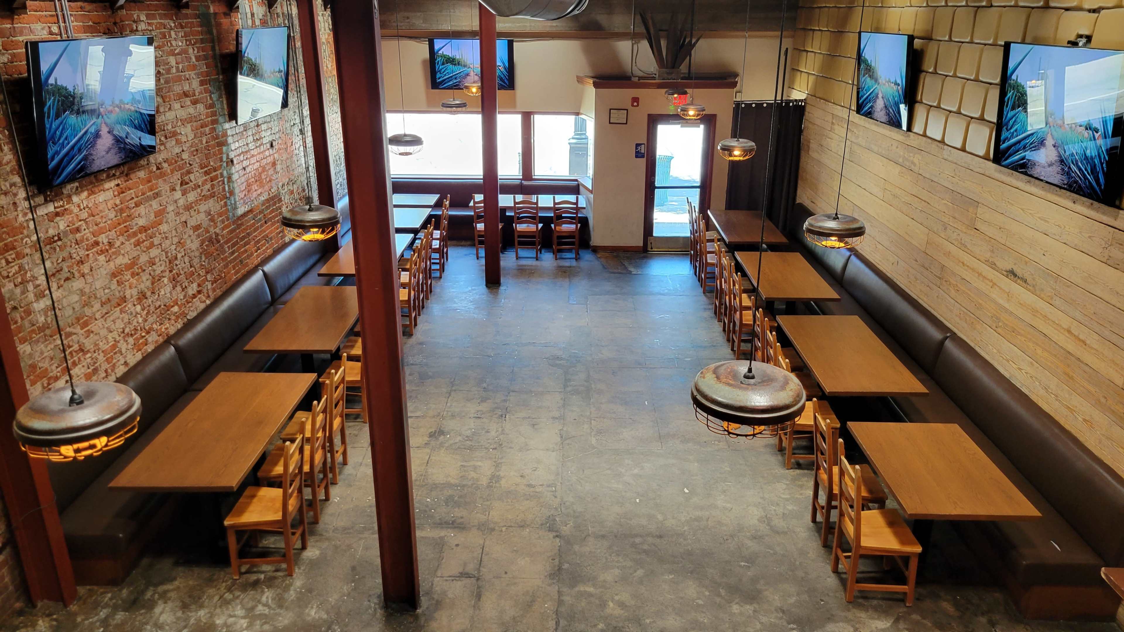 An indoor restaurant setting with wooden tables arranged in rows, brick and wooden walls, and multiple televisions mounted on the walls.