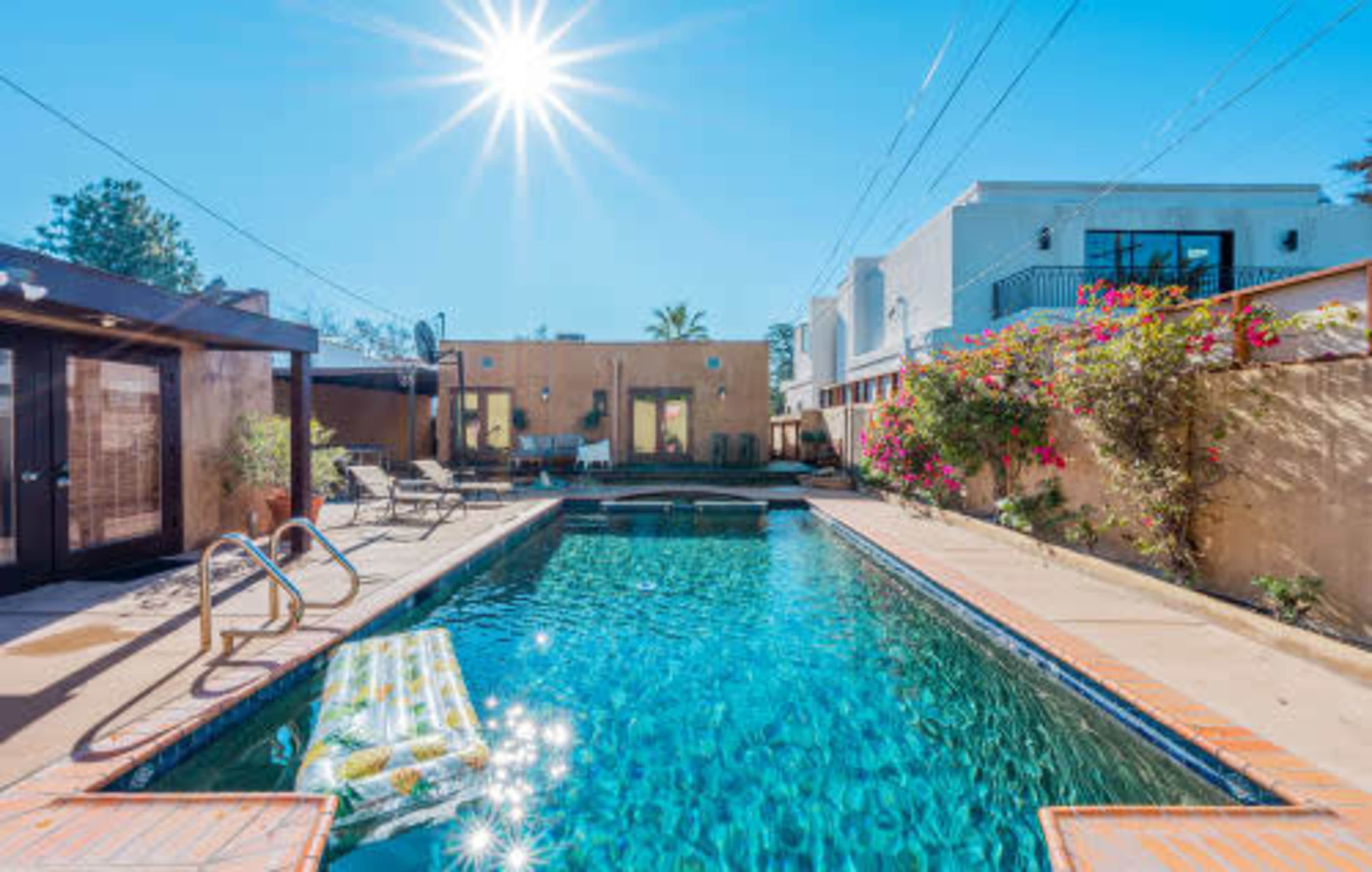A clear blue swimming pool is surrounded by lounge chairs and vibrant flowers, with buildings in the background under a bright sun.