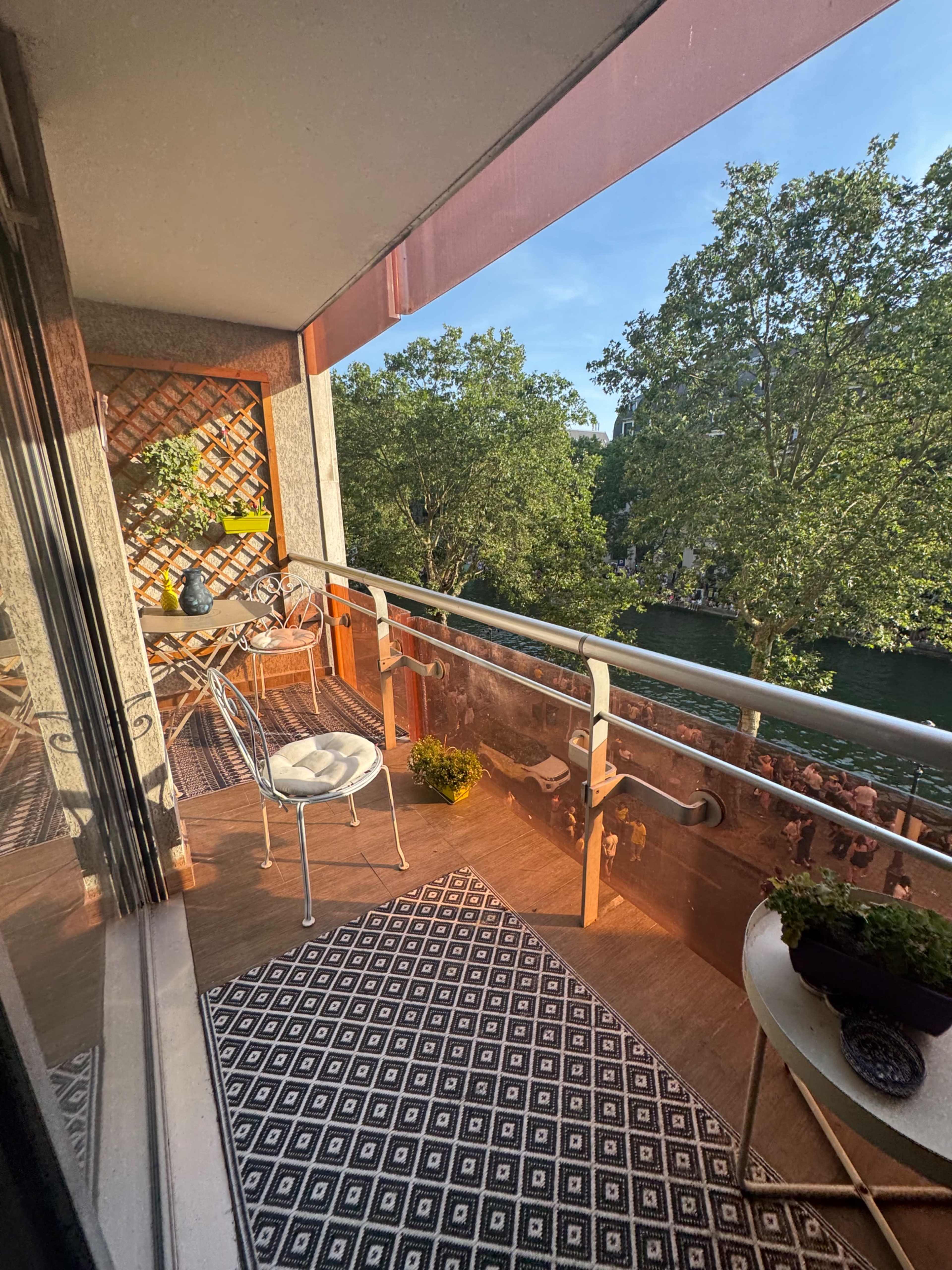 A small balcony features a patterned rug, a table and chair set, and potted plants, with trees visible outside.