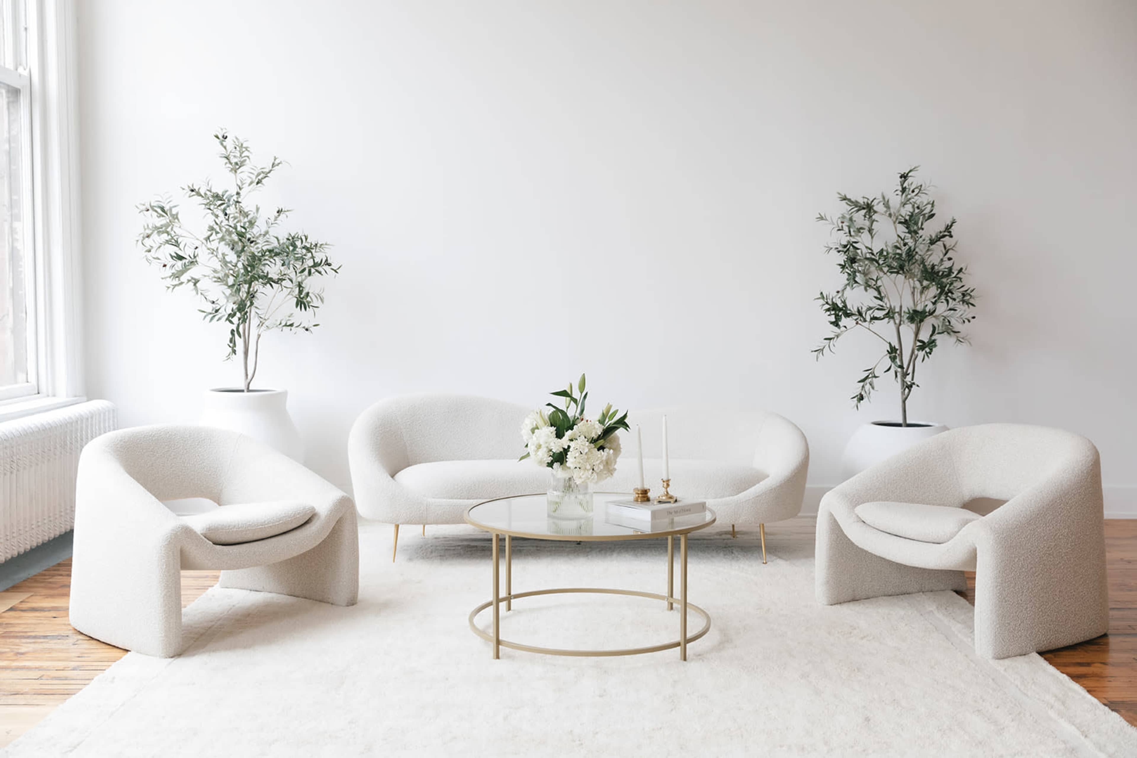 A bright, minimalistic living room featuring two white, curved sofas and two matching armchairs arranged around a glass coffee table with a floral centerpiece.