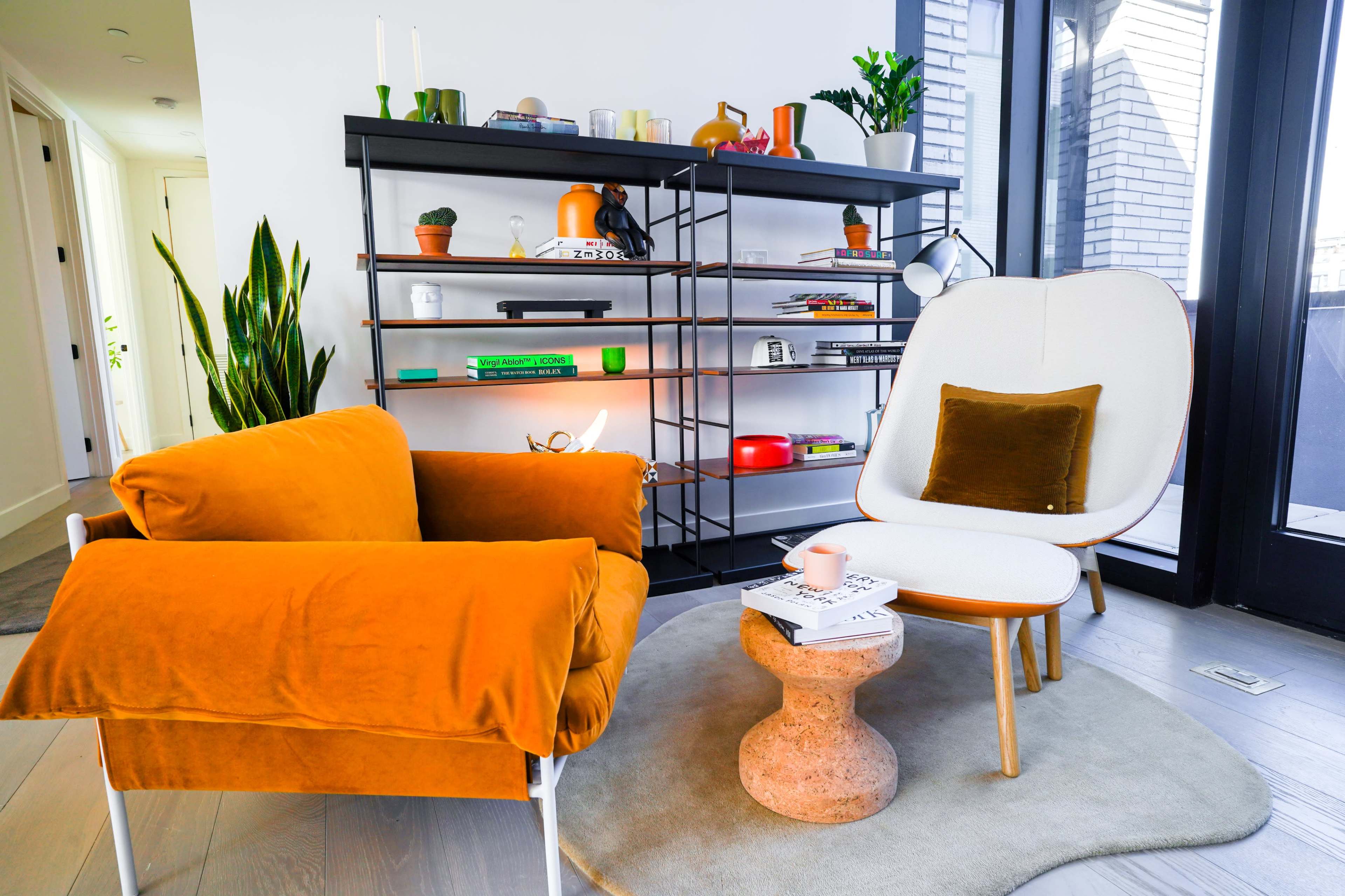 The scene features an orange velvet chair next to a light-colored textured side table, with a modern bookshelf displaying various decorative items and books in a well-lit room.
