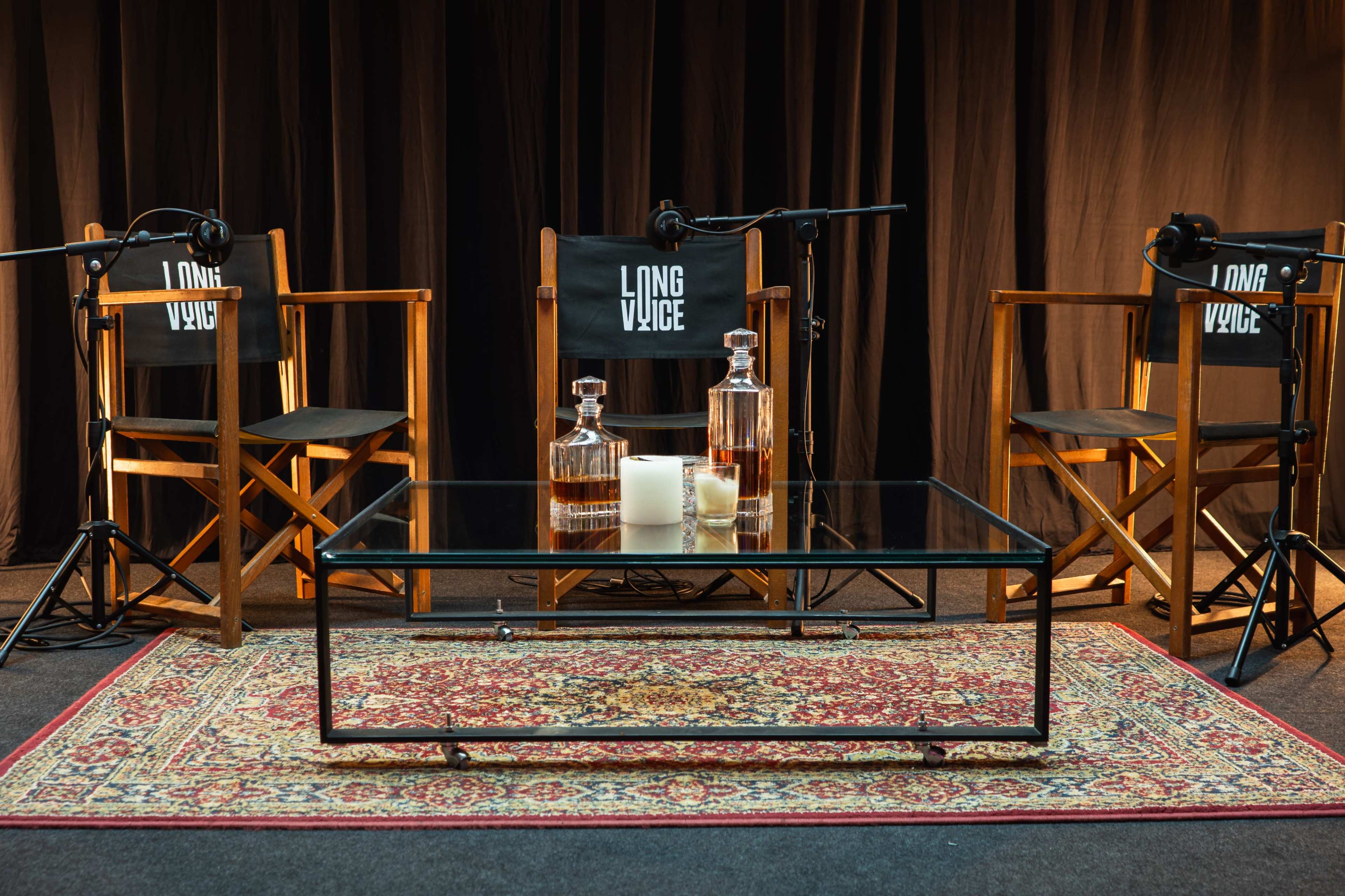 The image shows a set designed for an interview or podcast, featuring three empty director's chairs, a glass coffee table with two decanters and a candle, all arranged on a decorative rug in front of a dark backdrop.