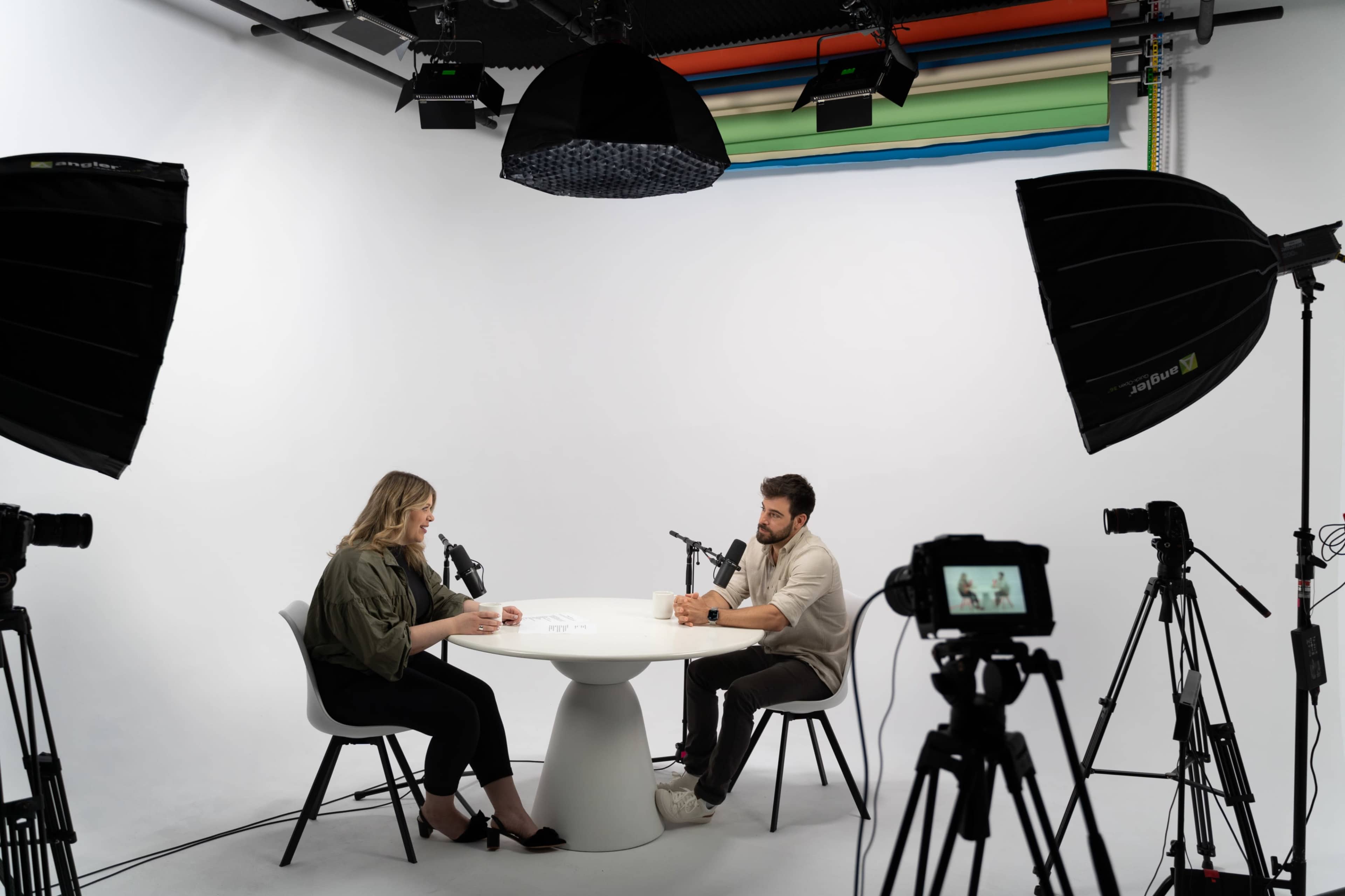 A man and a woman sit at a round table in a studio, each speaking into a microphone while multiple cameras are positioned around them.