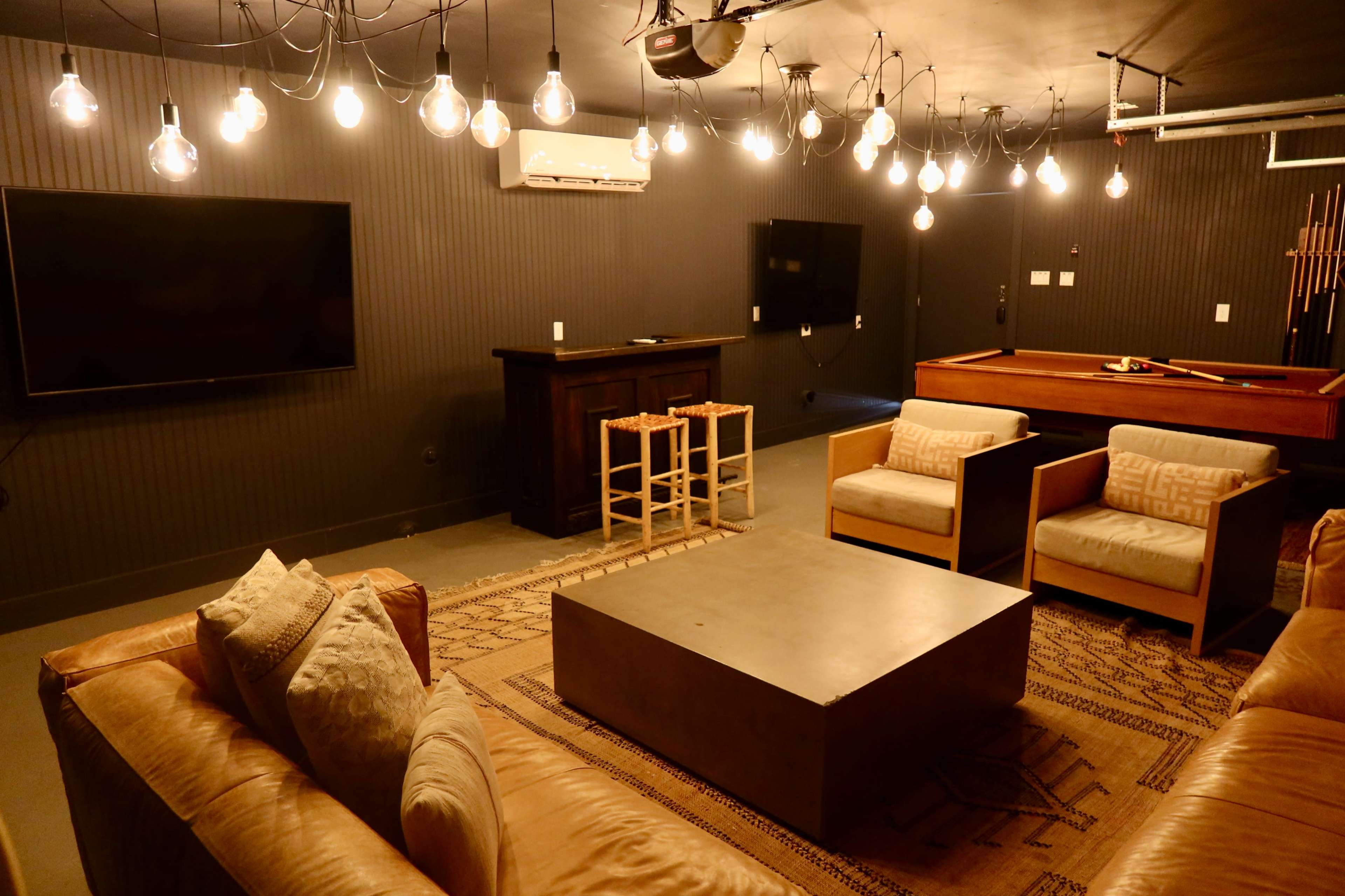 The image shows a modern lounge area with a sectional sofa, a coffee table, two chairs, a pool table, and multiple hanging light bulbs.