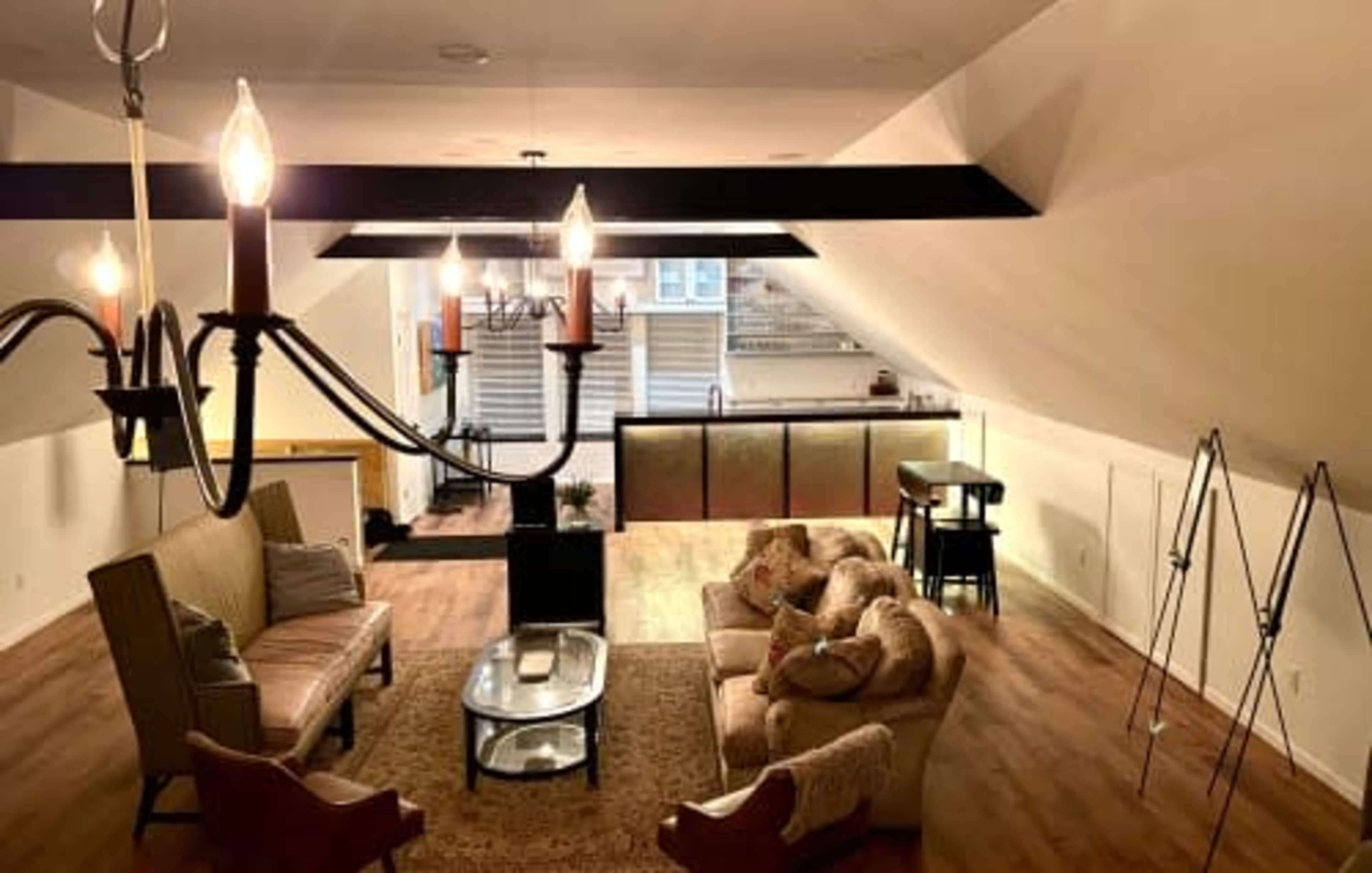 A spacious attic living area with a chandelier, a sectional sofa, a coffee table, and a kitchenette visible in the background.