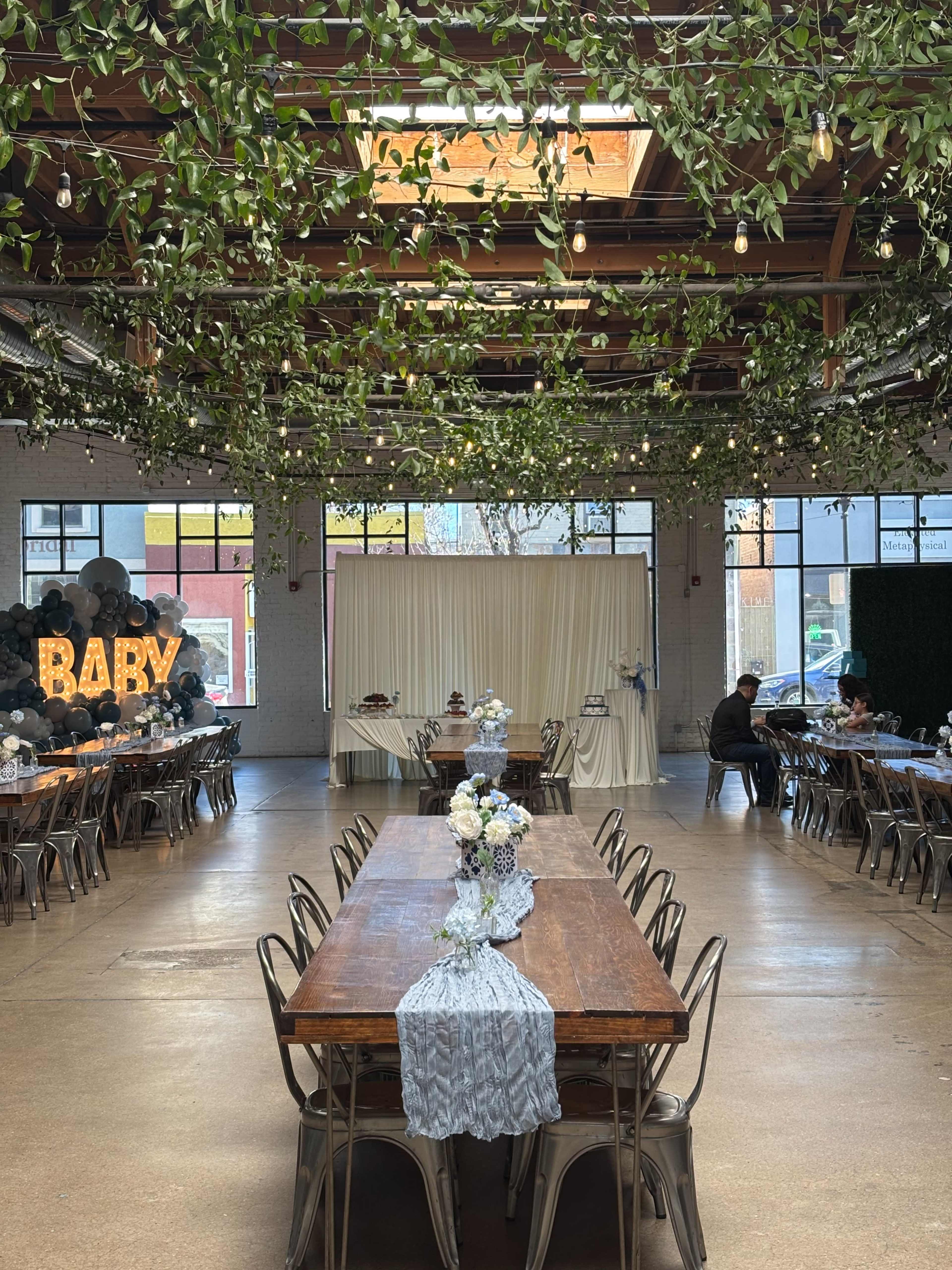 The image shows a spacious event hall with long wooden tables adorned with centerpieces, decorated with greenery overhead, and a backdrop featuring a "BABY" sign.