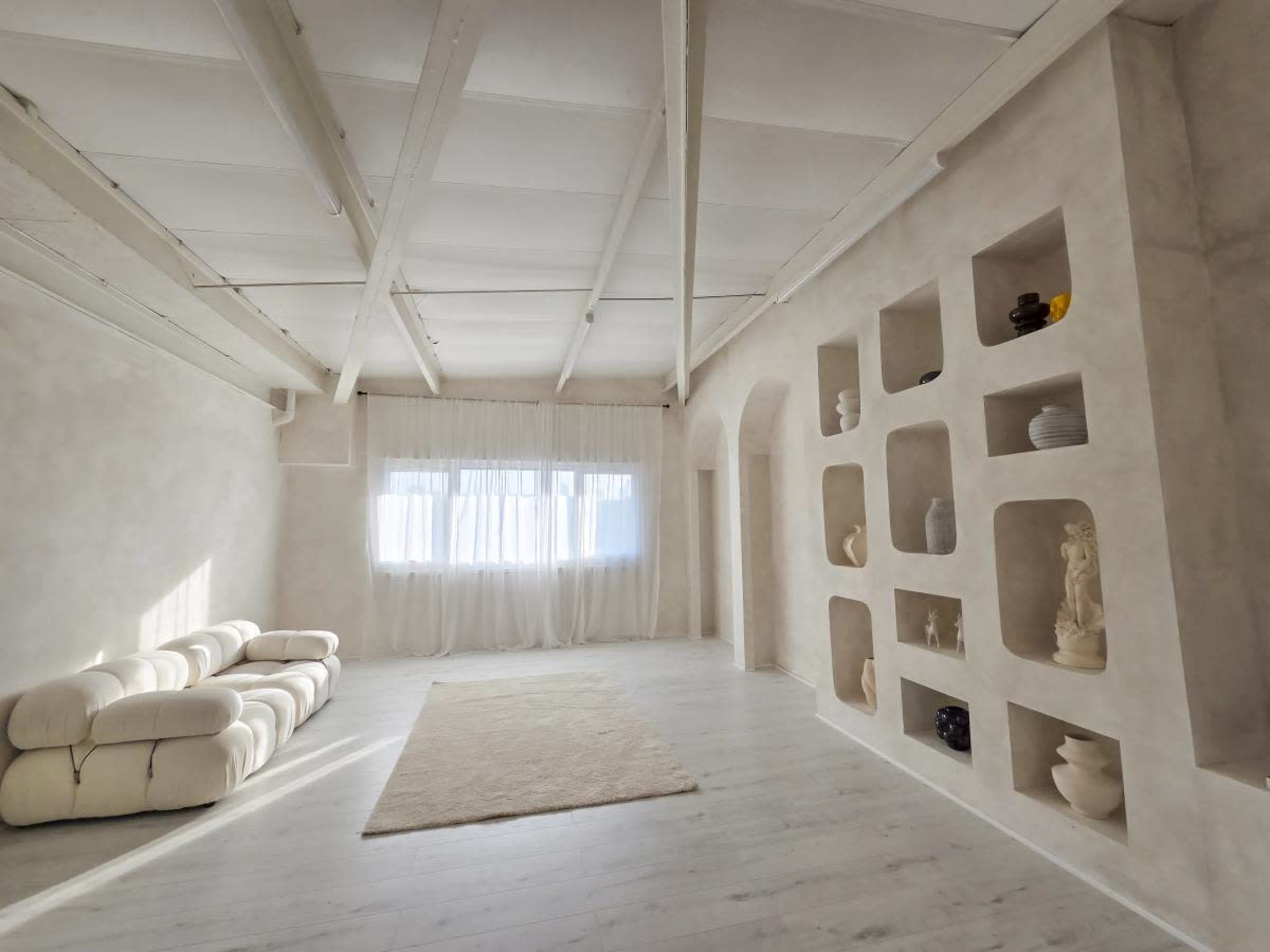 The image shows a spacious, minimalist room with a white color scheme, featuring a plush sofa, large windows with sheer curtains, and a wall display of various decorative objects.