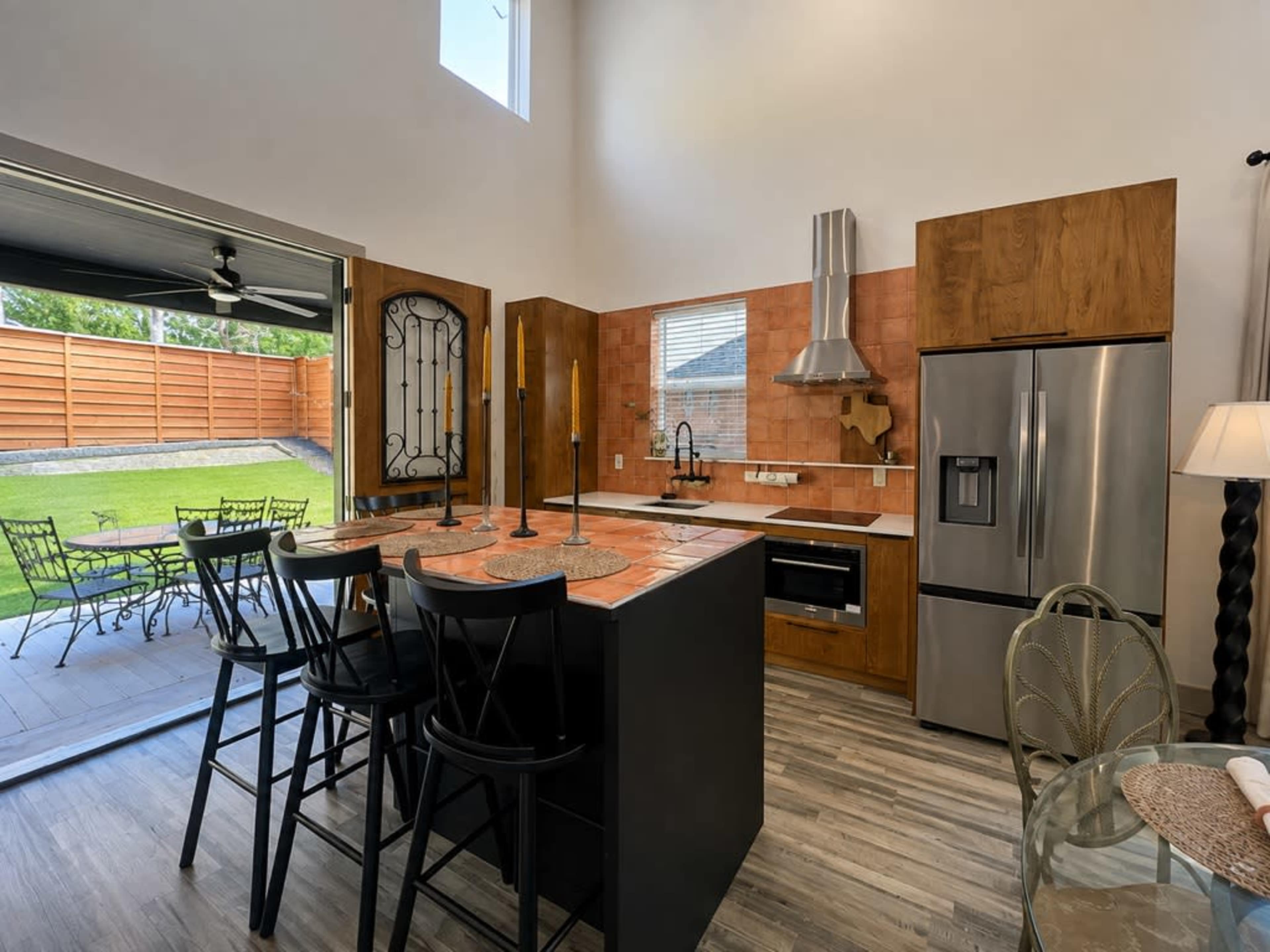 The image shows a modern kitchen with an open layout, featuring a central island with seating, stainless steel appliances, and a view of an outdoor patio through large sliding doors.
