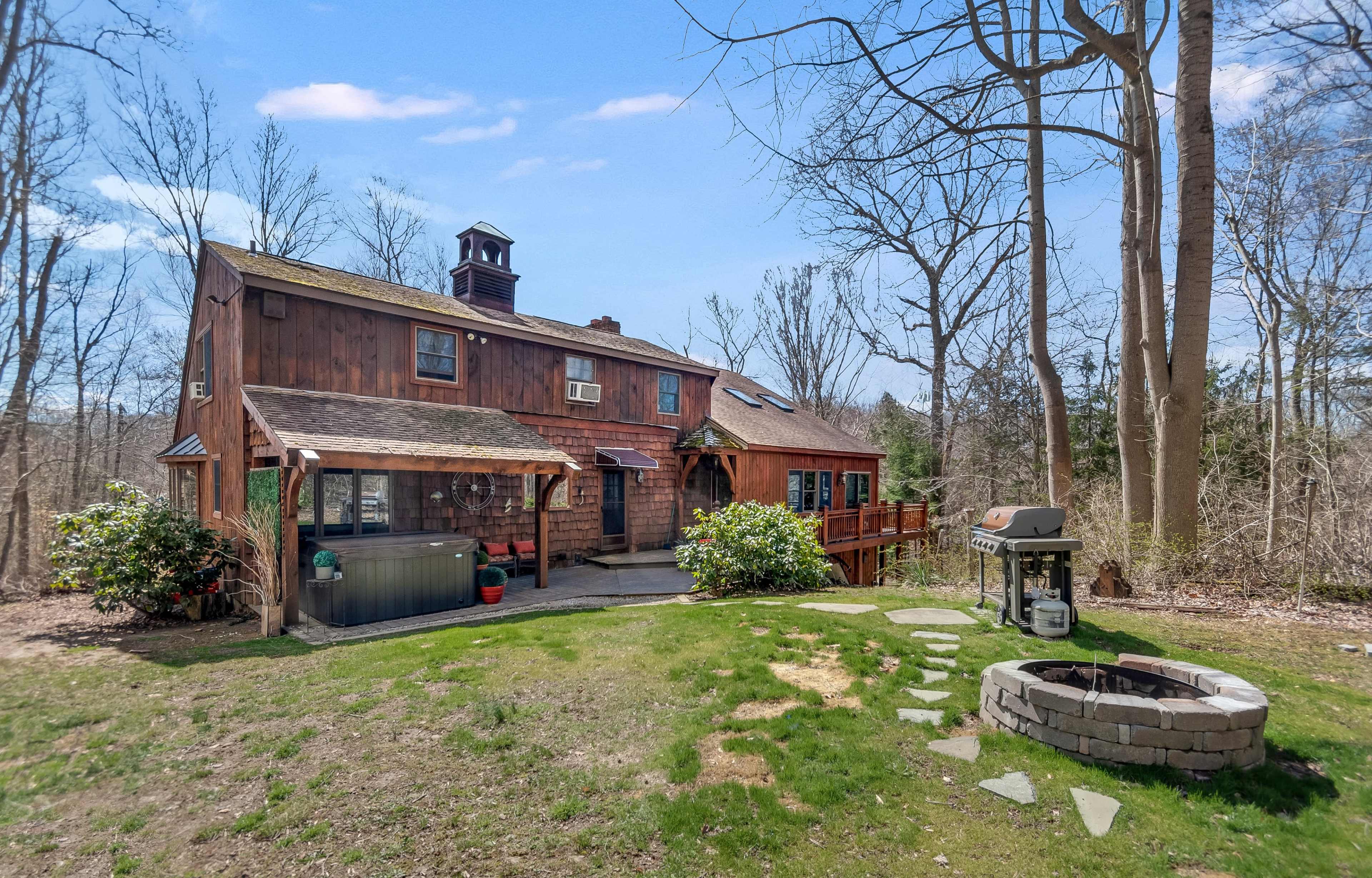 A wooden house with a deck and a stone fire pit sits surrounded by trees in a grassy area.