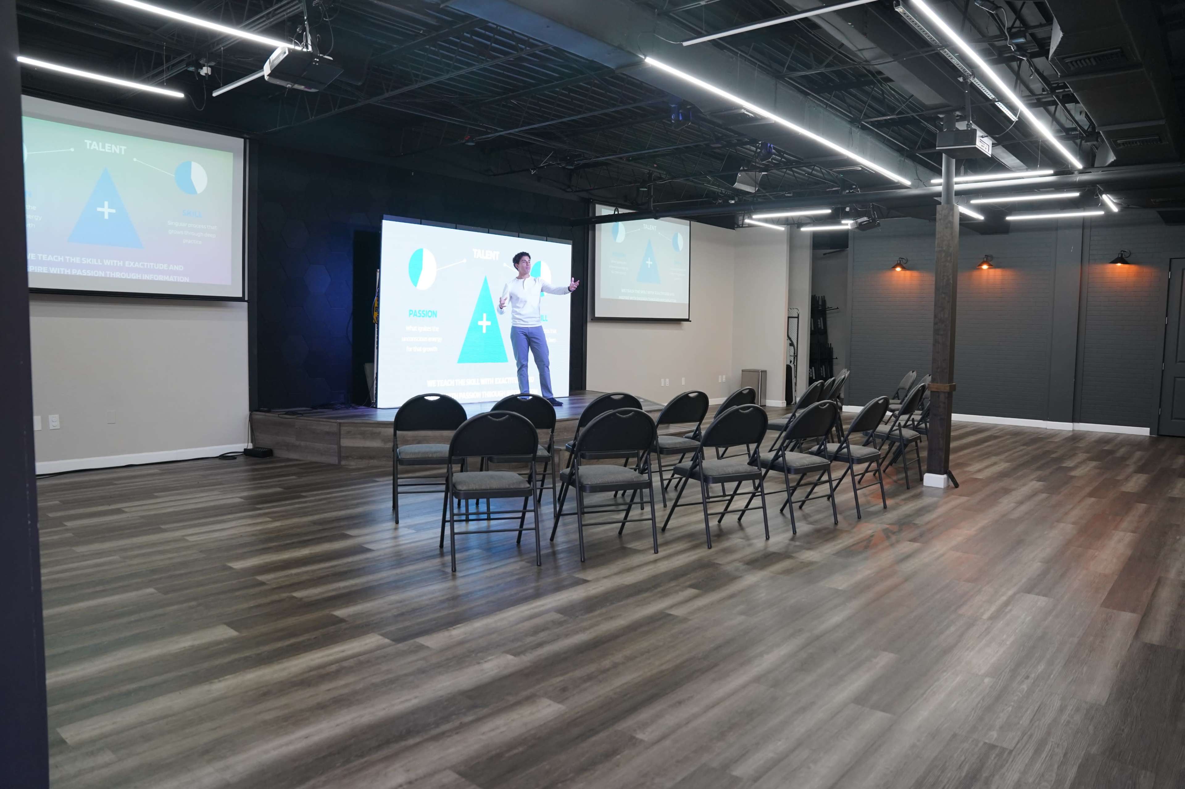 A presenter stands on a stage in a light-filled conference room with chairs arranged in rows facing large projection screens displaying graphics.