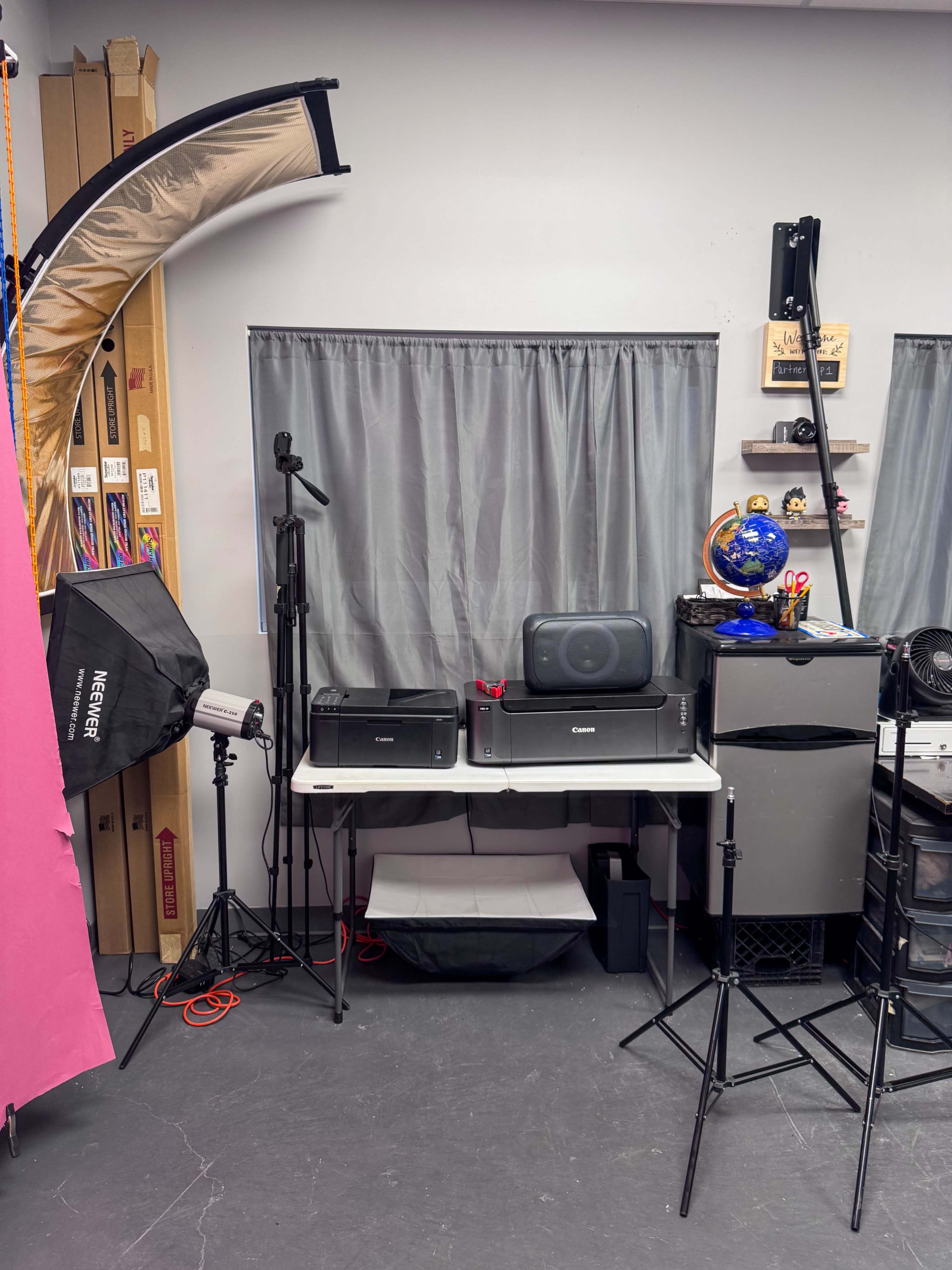 The image shows a workspace featuring two printers, a monitor, a small fridge, and various photographic equipment against a backdrop of gray curtains and organized shelves.