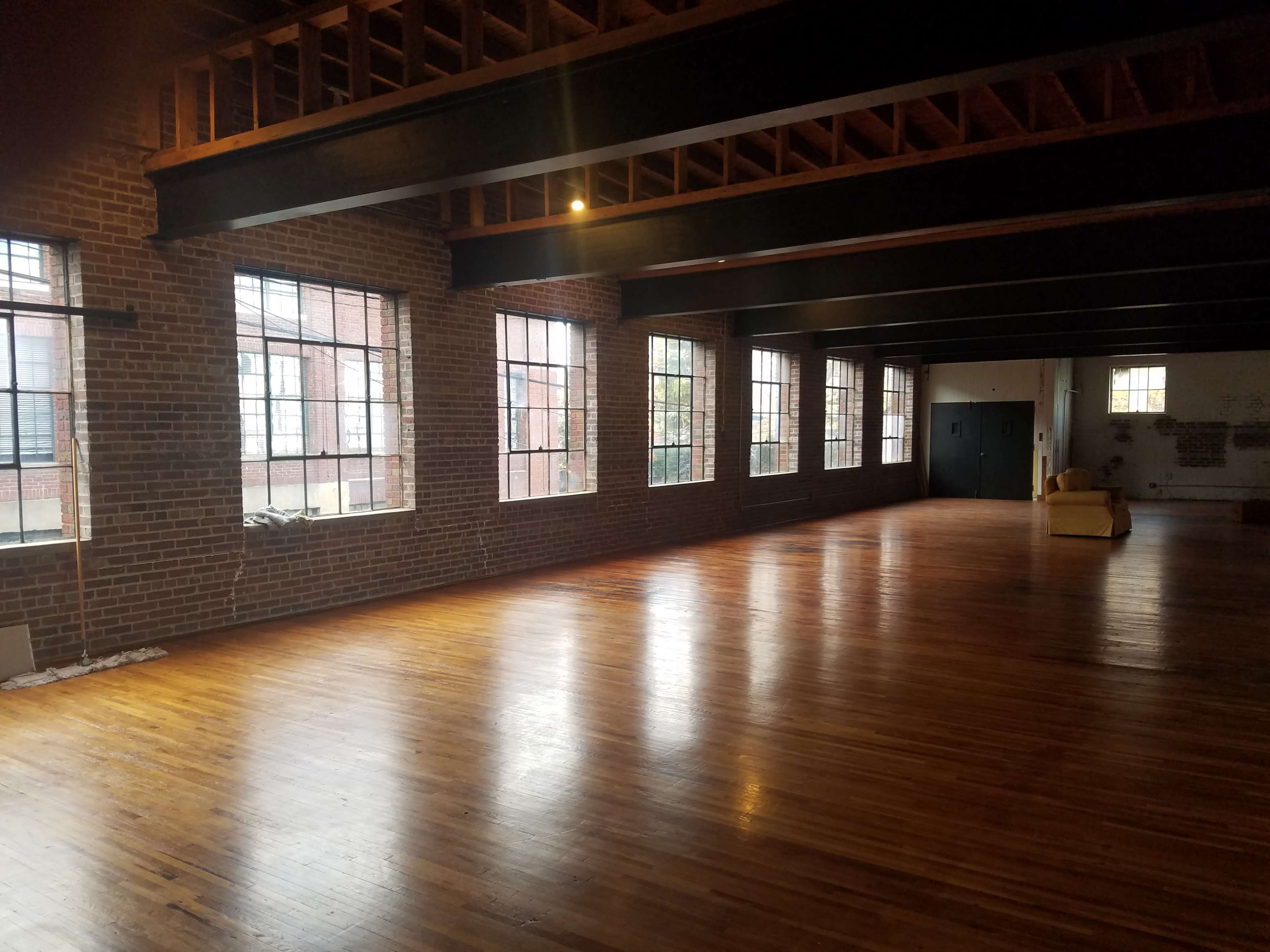 A spacious, empty room with wooden floors and large windows along the brick walls.