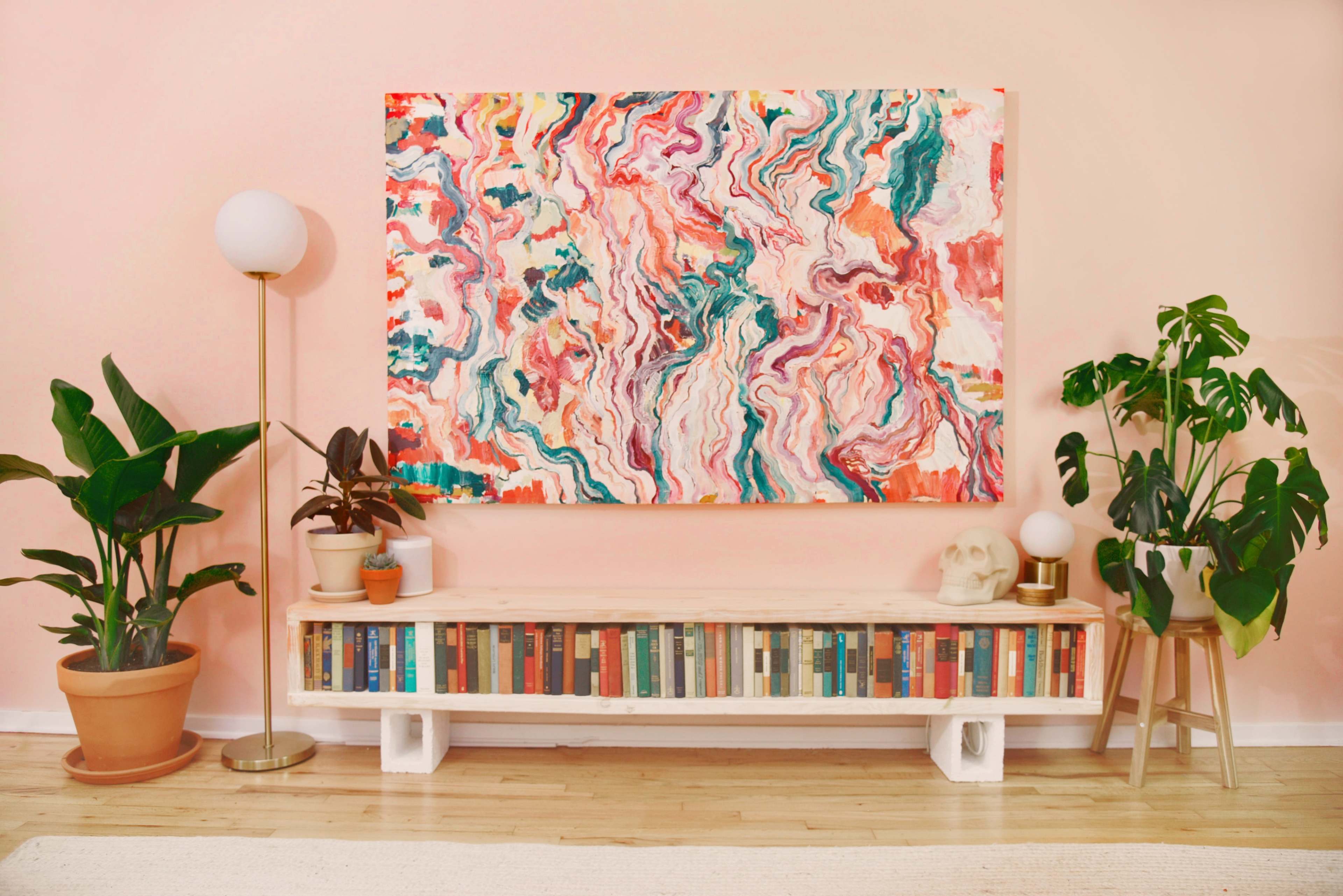 A brightly colored abstract painting hangs above a wooden shelf filled with books, flanked by potted plants and two lamps on either side.