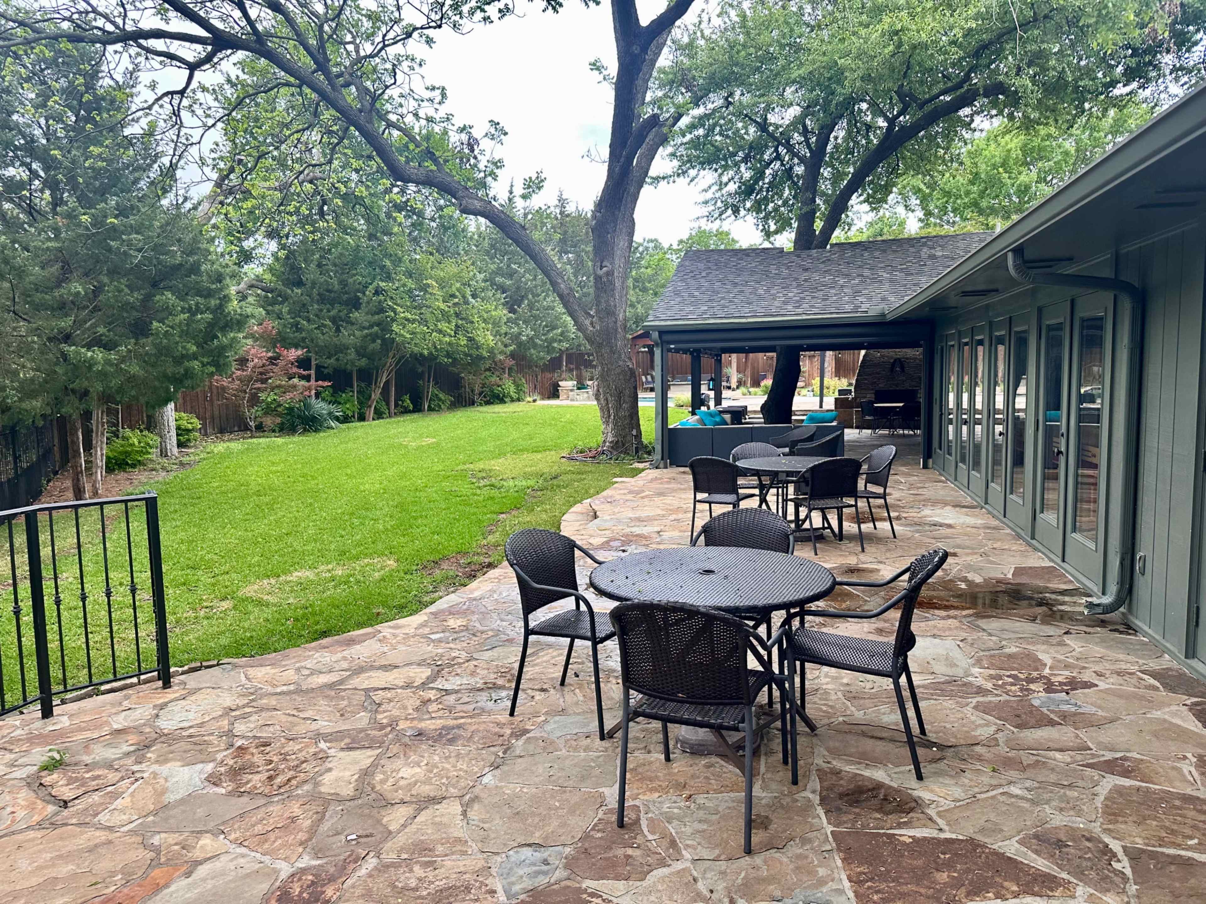 A stone patio with several metal tables and chairs overlooks a green lawn surrounded by trees.