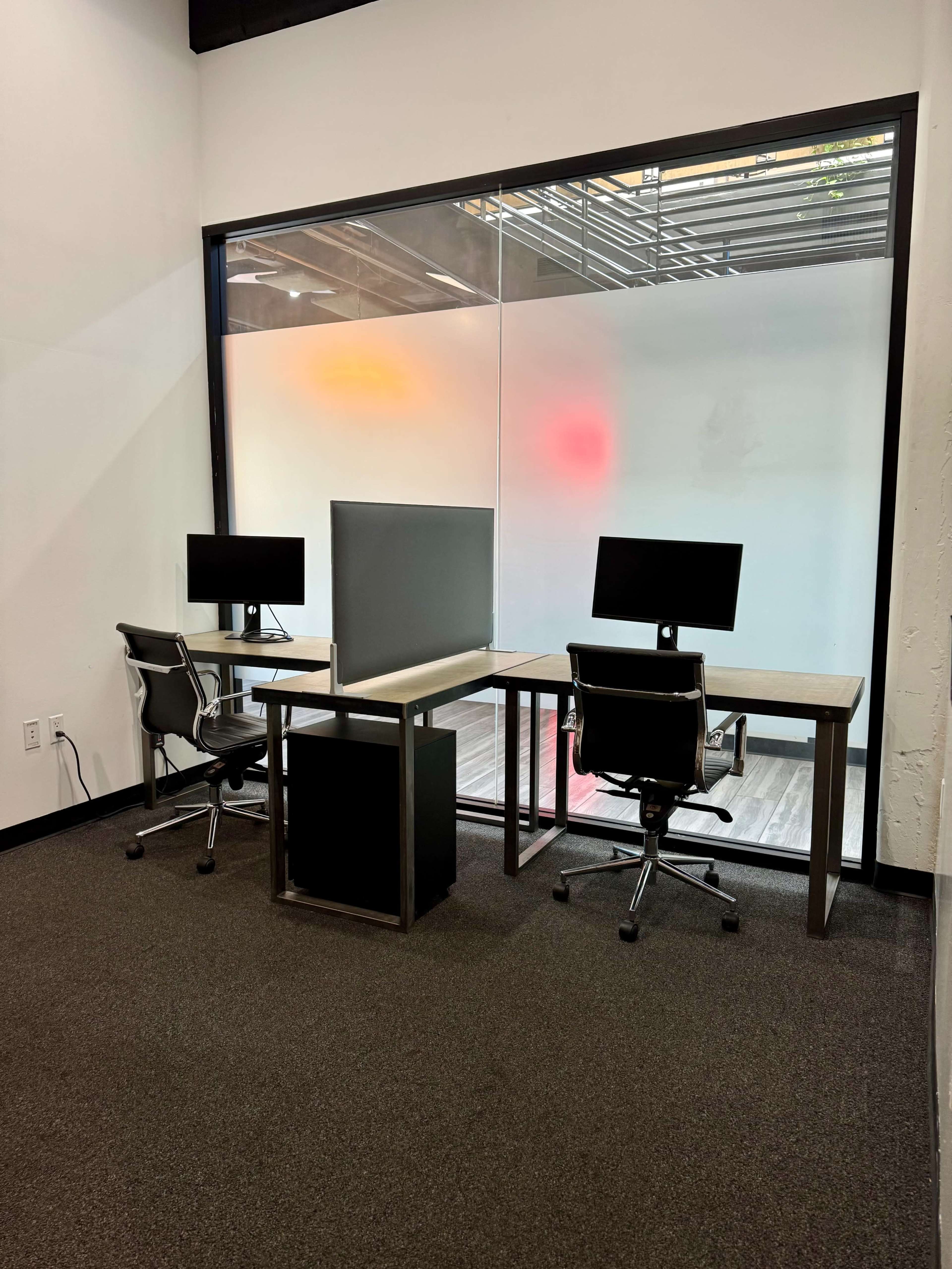The image shows a minimalist office space with two desks, each equipped with a computer, facing a large window with colorful designs.