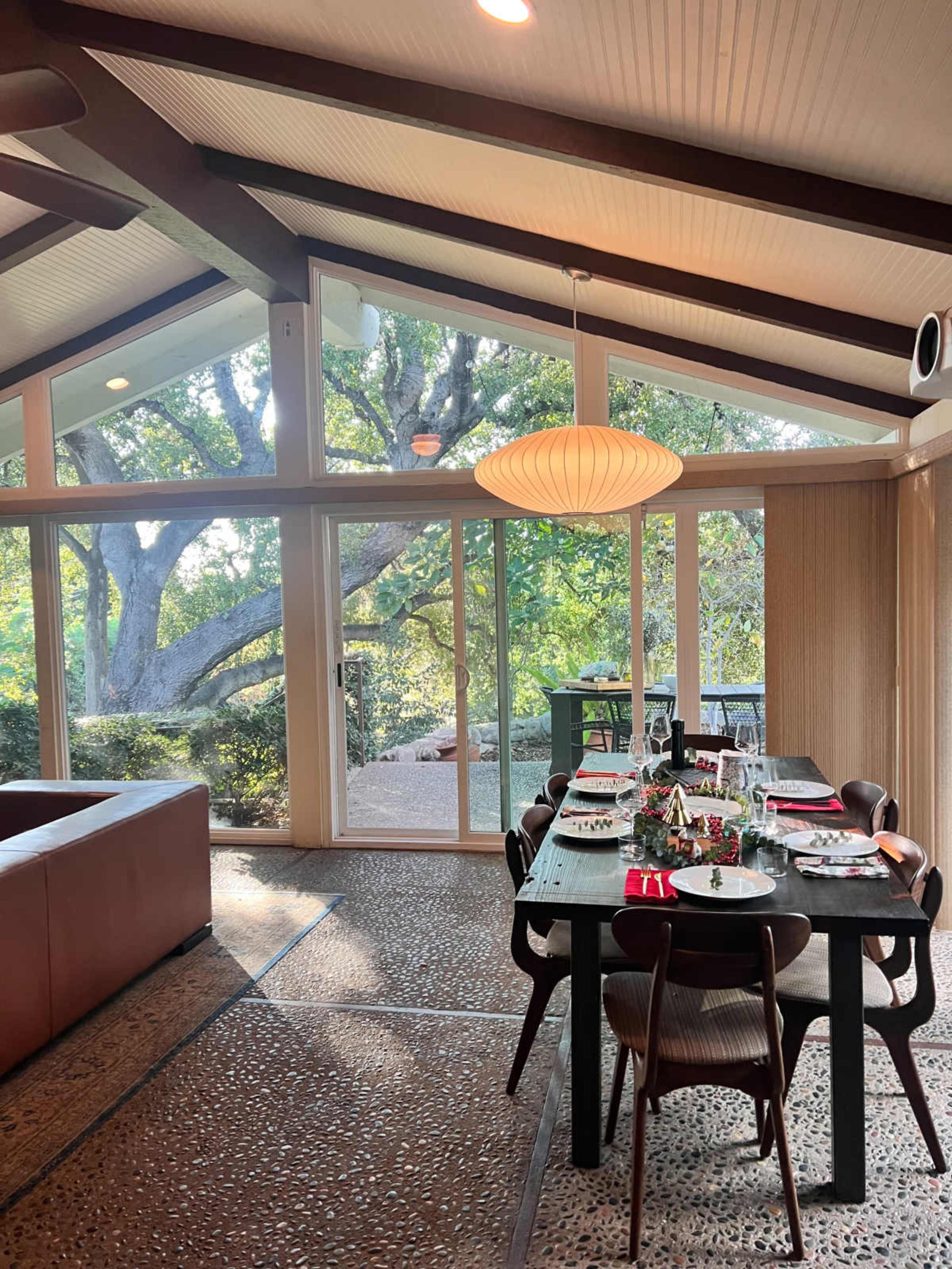The image shows a modern dining area with a large table set for a meal, featuring a view of trees outside through large glass windows.