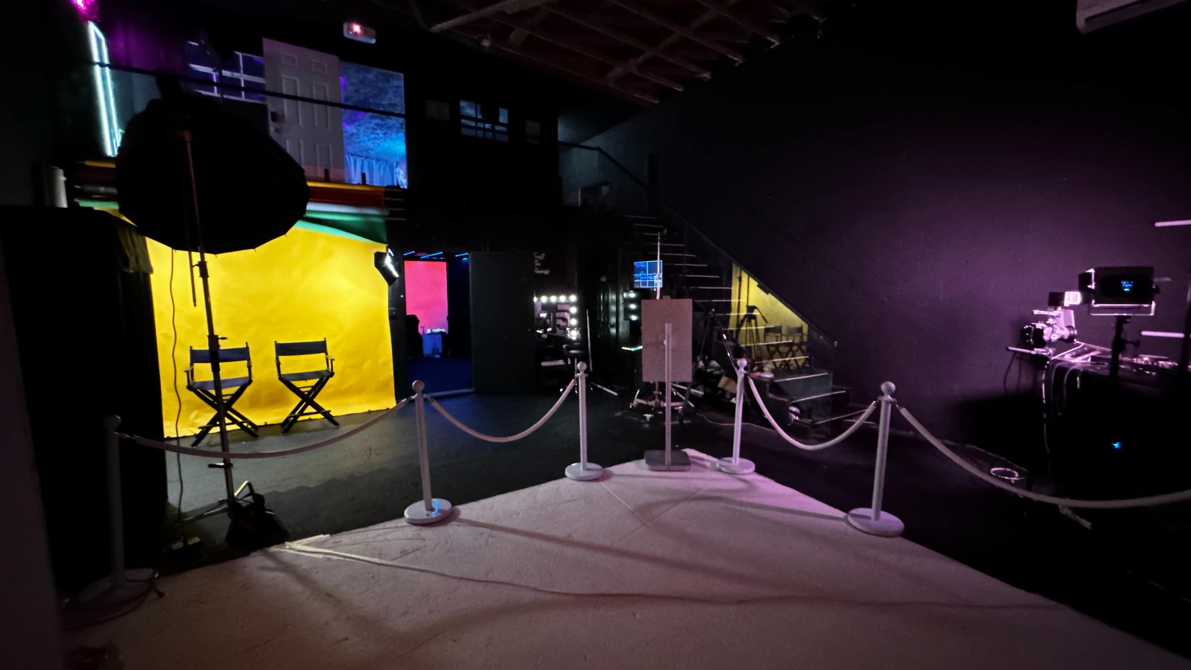 The image shows a studio space with colorful backdrops, lighting equipment, and a pathway marked by a rope barrier leading to a staircase.