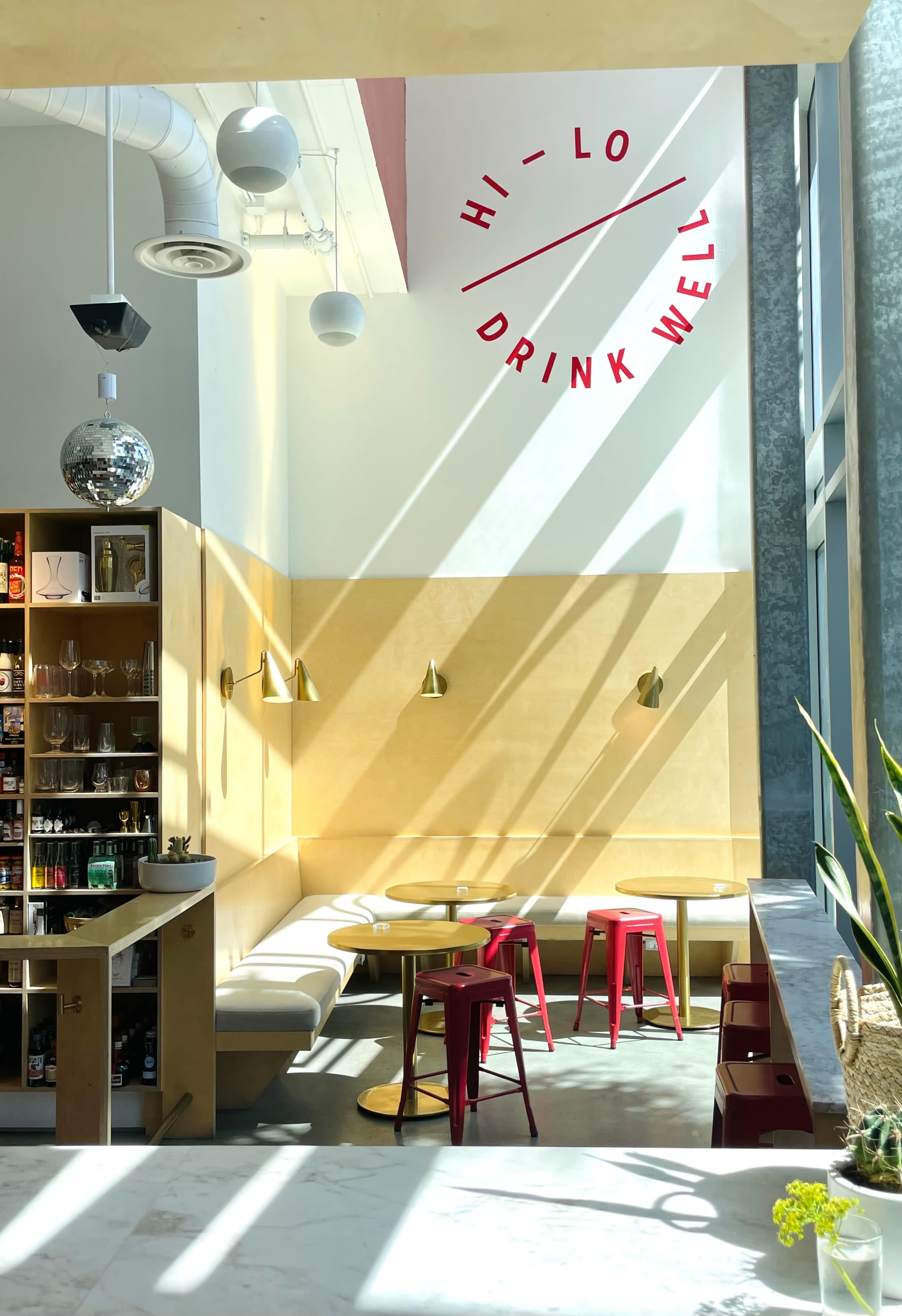 A brightly lit café features light wooden seating, red tables, and a decorative wall with the phrase "HI - LO DRINK WELL."