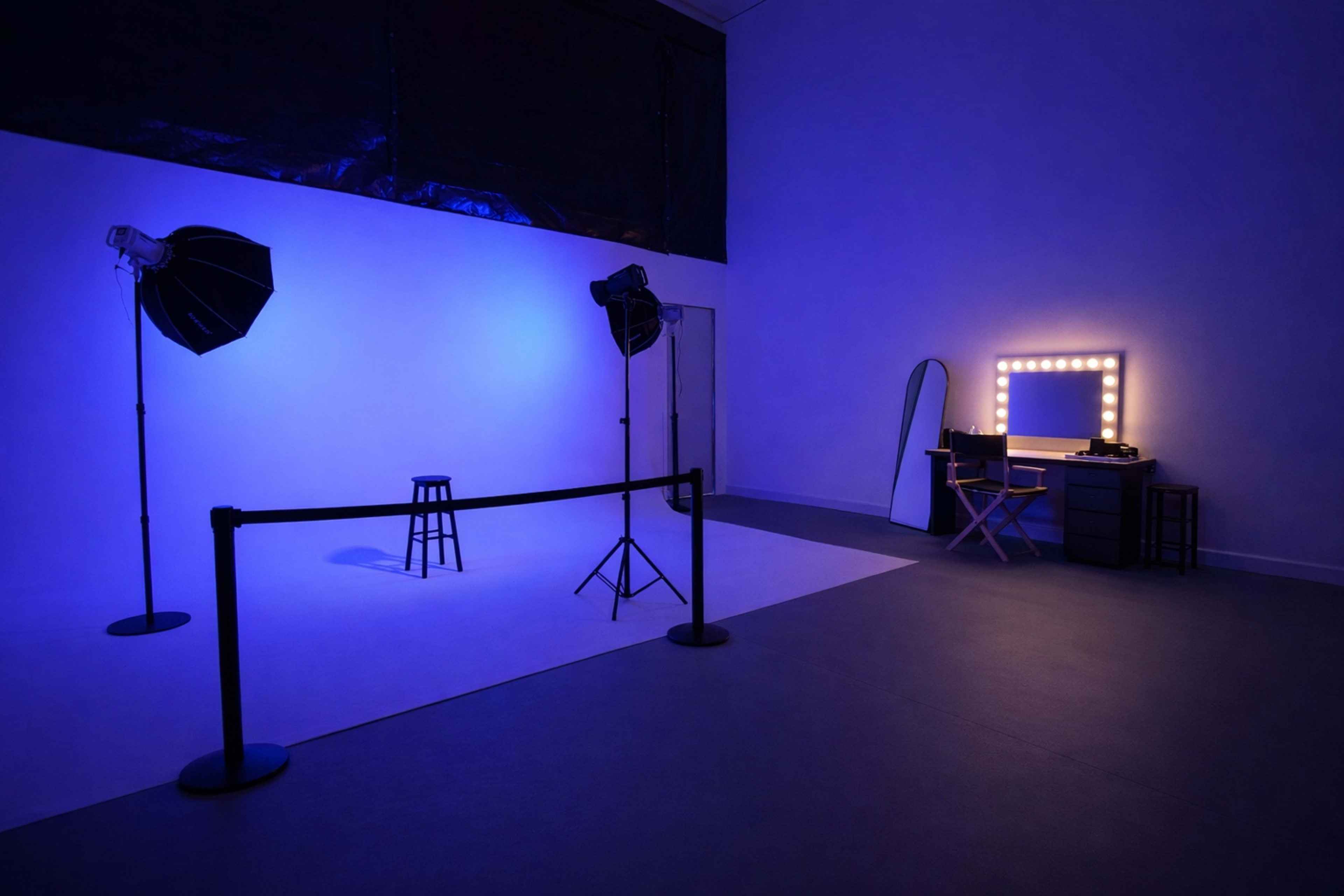The image shows a photography studio with blue lighting, featuring two softbox lights, a stool, a makeup mirror with lights, and a director's chair.